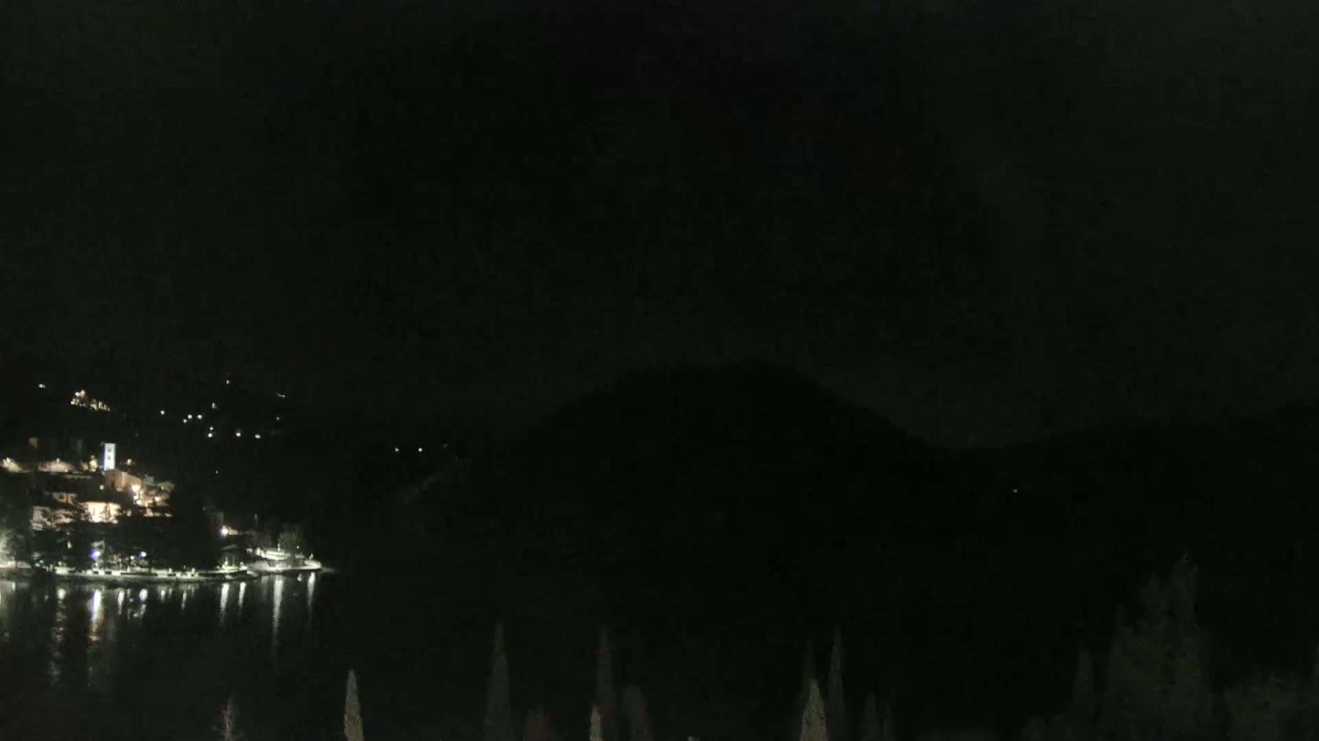 A nighttime view shows a dark, calm body of water reflecting the lights of a town nestled against a dark mountain.