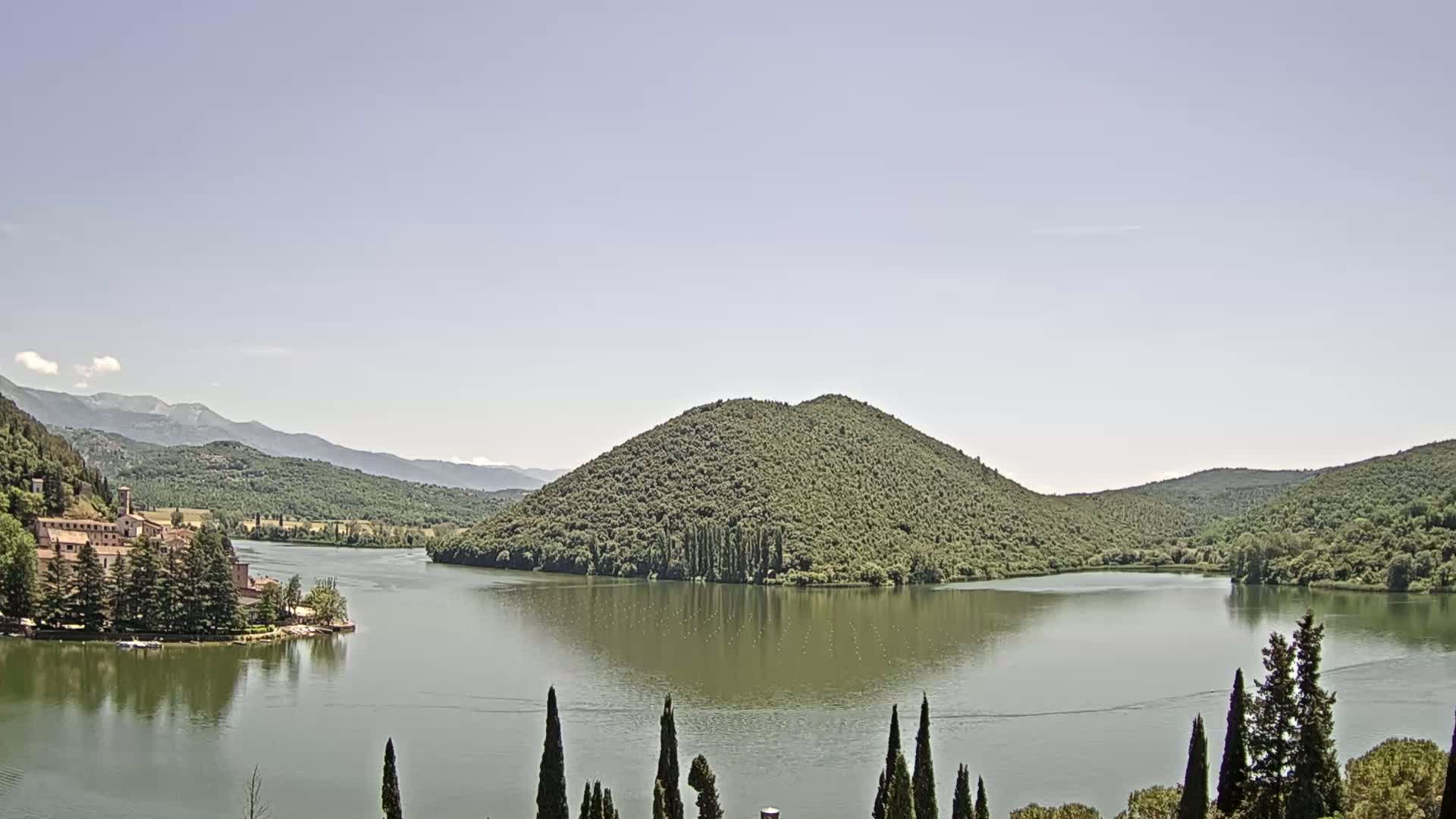 A calm lake is nestled between tree-covered hills and mountains under a clear, light-blue sky.