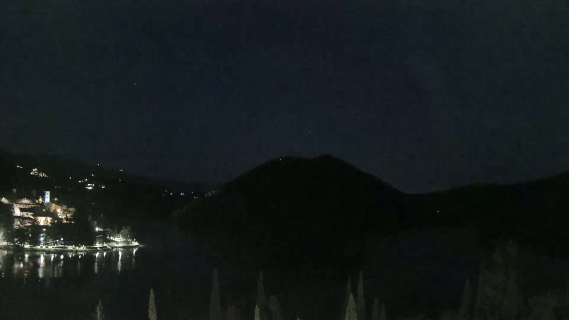 A nighttime view shows a dark lake reflecting the lights of a small town nestled among dark hills under a clear, starry sky.