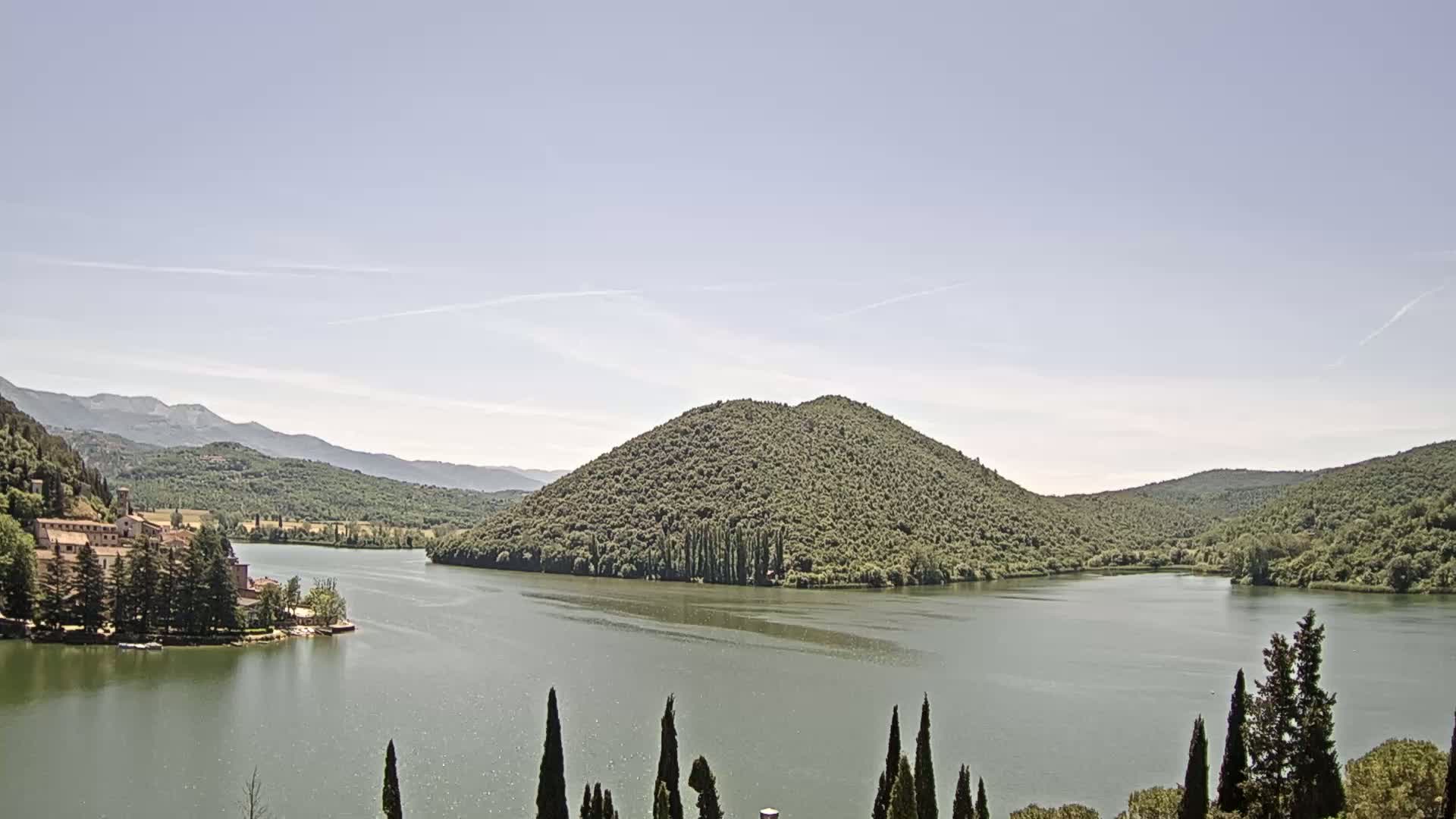 A calm lake is surrounded by lush green hills and mountains under a clear, light blue sky.