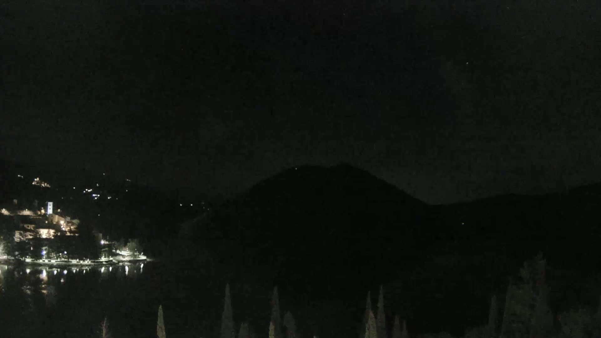 A nighttime view shows a dark, still lake reflecting lights from a village nestled on a hillside beneath a large, dark mountain.