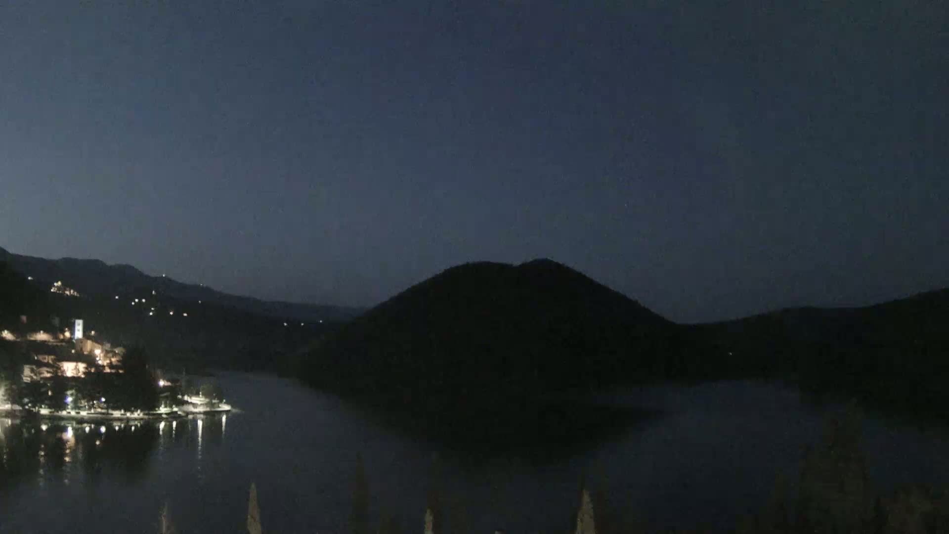 A dark, nighttime view shows a body of water reflecting the lights of a small town nestled beside hills under a twilight sky.