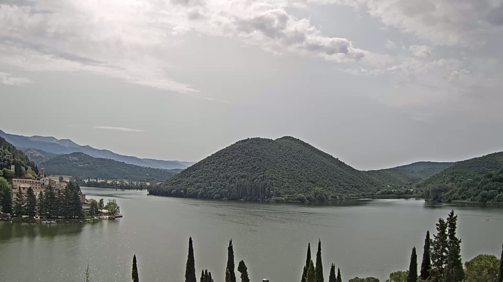 A calm lake surrounded by green hills and a building complex on the left shore under a mostly cloudy sky.