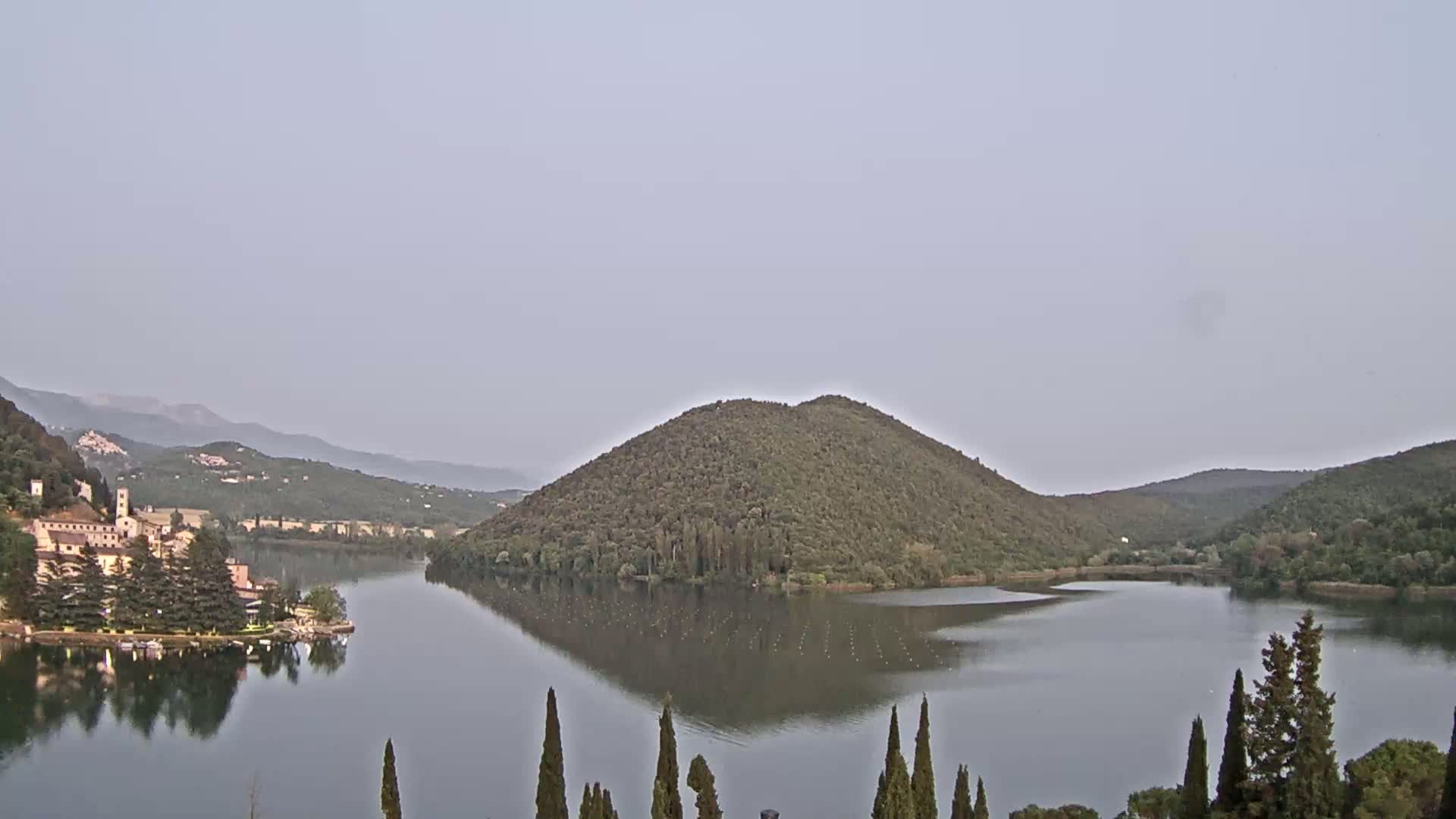 A calm lake is nestled between tree-covered hills under a hazy sky.