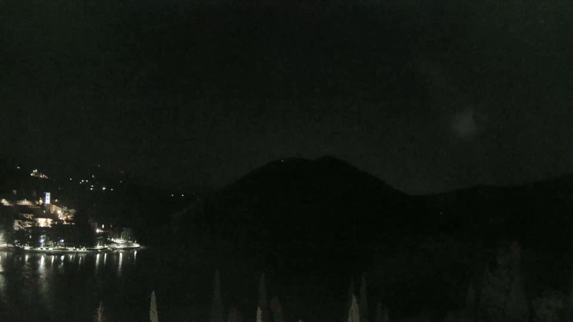 A nighttime view of a dark, silhouetted mountain overlooking a calm body of water with a shoreline featuring sparsely lit buildings.