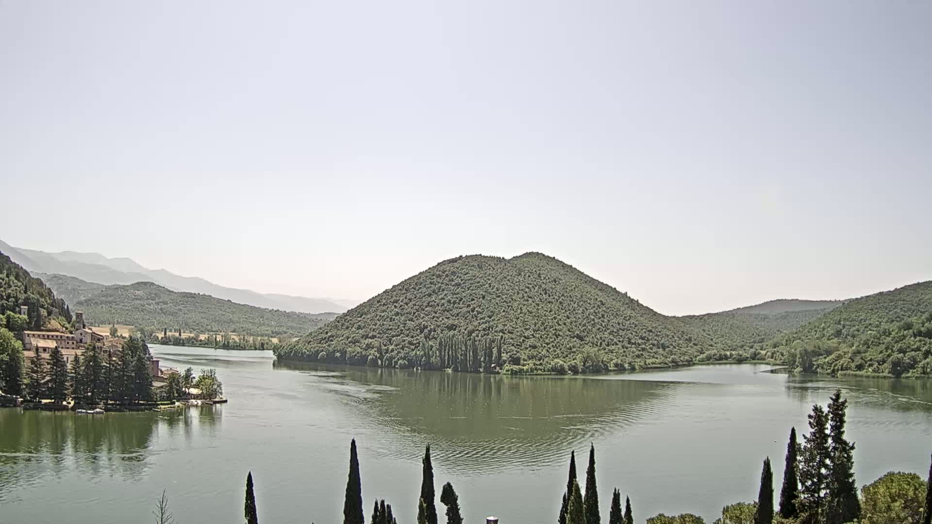 A calm lake is nestled within a valley of green hills and mountains under a bright, clear sky.