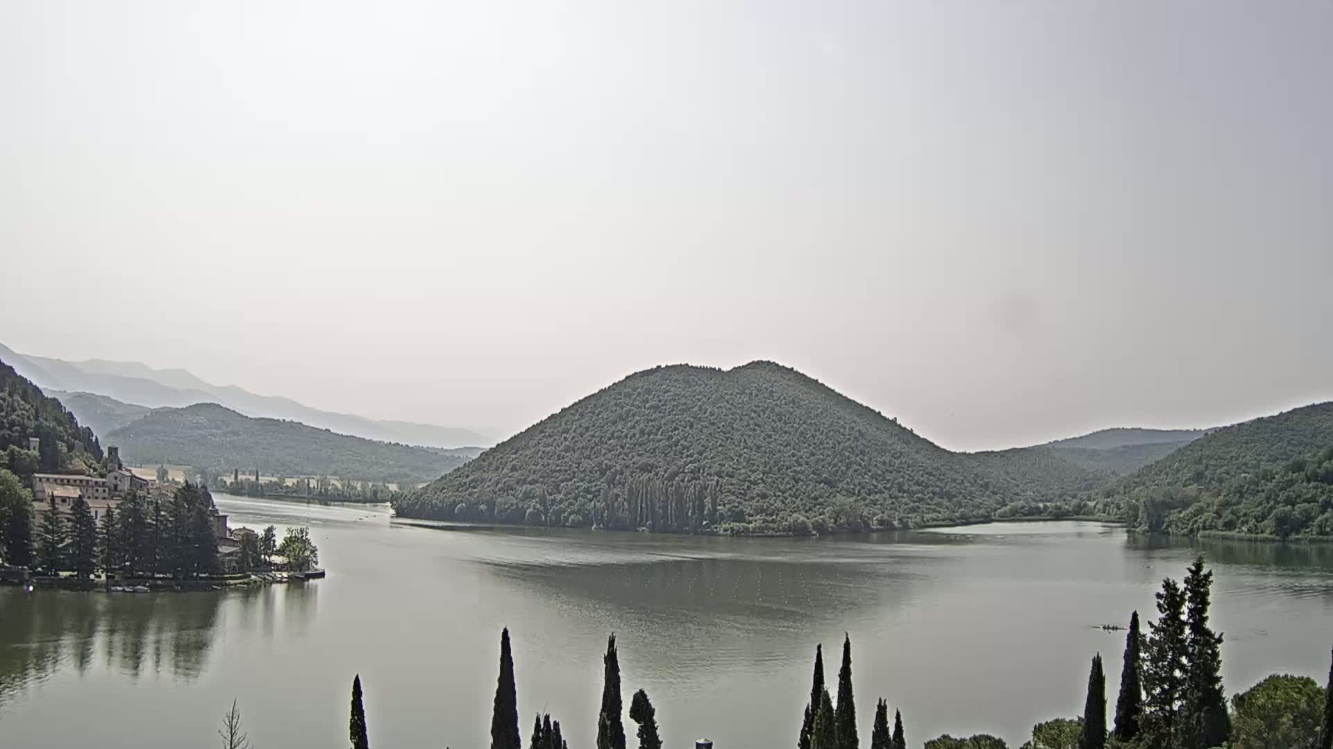 A calm lake is surrounded by green hills and mountains under a hazy sky, with a small building visible on the lakeshore.
