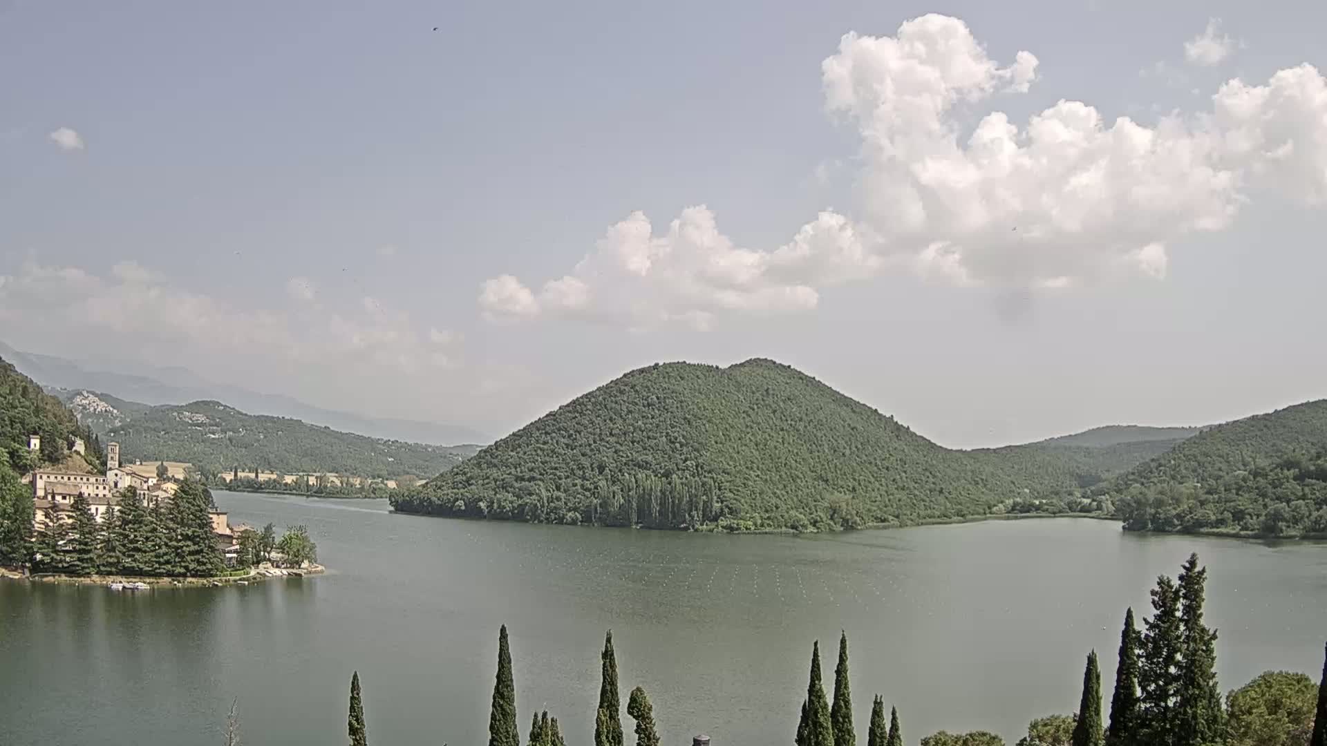 A tranquil lake is surrounded by lush green hills and mountains under a partly cloudy sky.