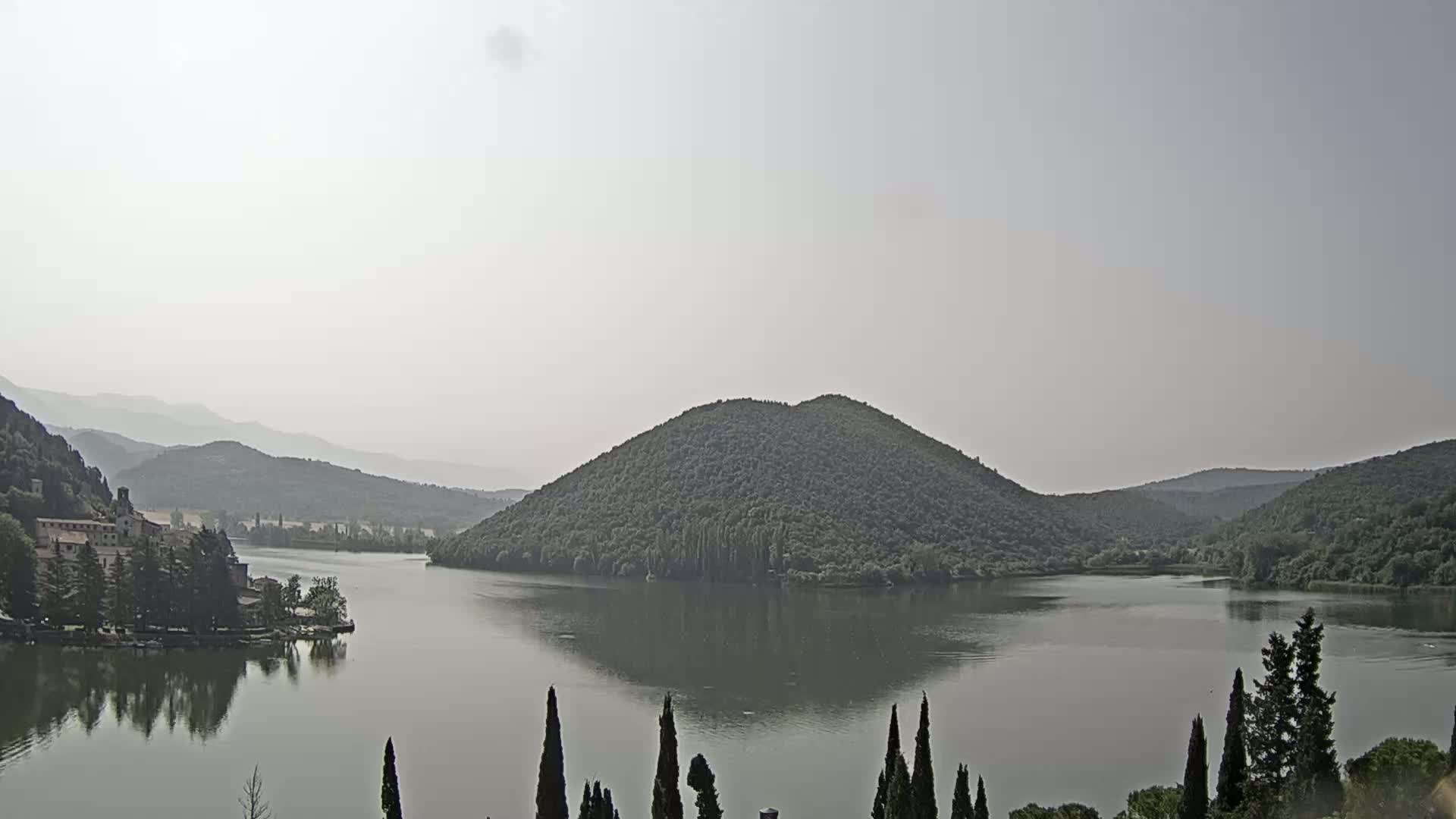 A calm lake reflects hazy mountains and a tree-covered island under a mostly clear sky.