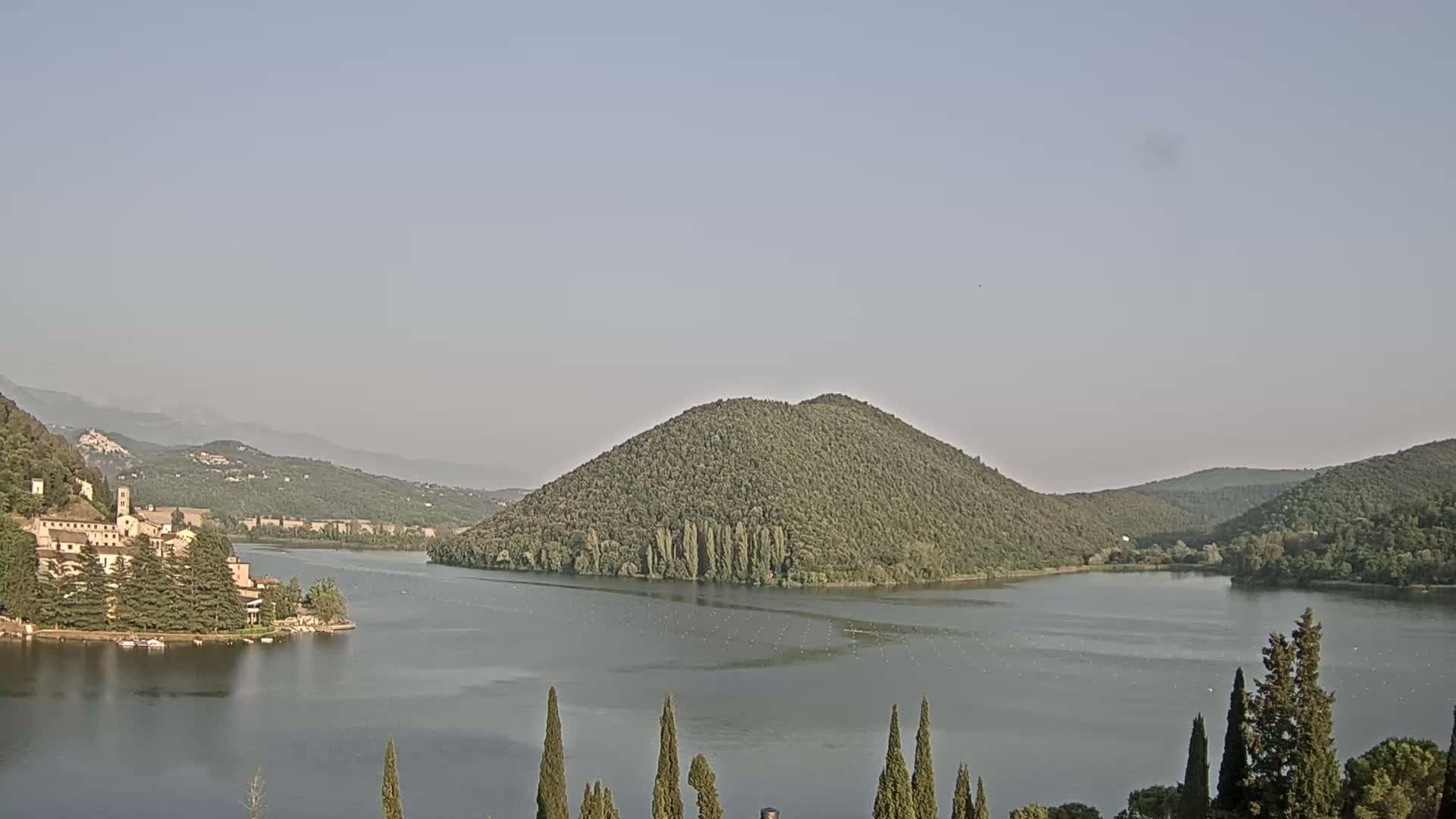 A calm lake is nestled between tree-covered hills and mountains under a clear, light-grey sky, with a village visible on the lakeshore.