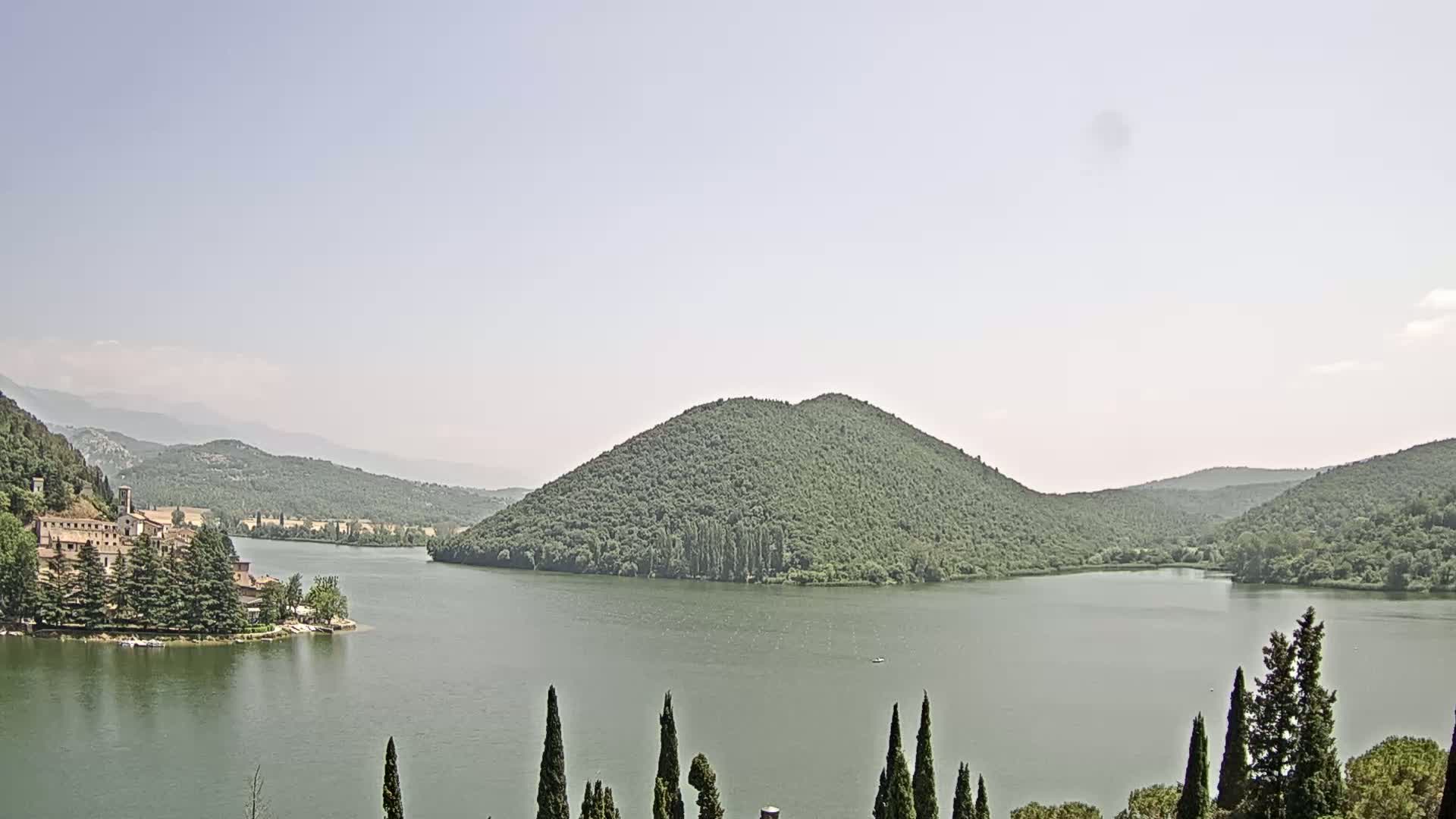 A calm lake is nestled between green hills and mountains under a hazy sky, with a building complex visible on a peninsula to the left.