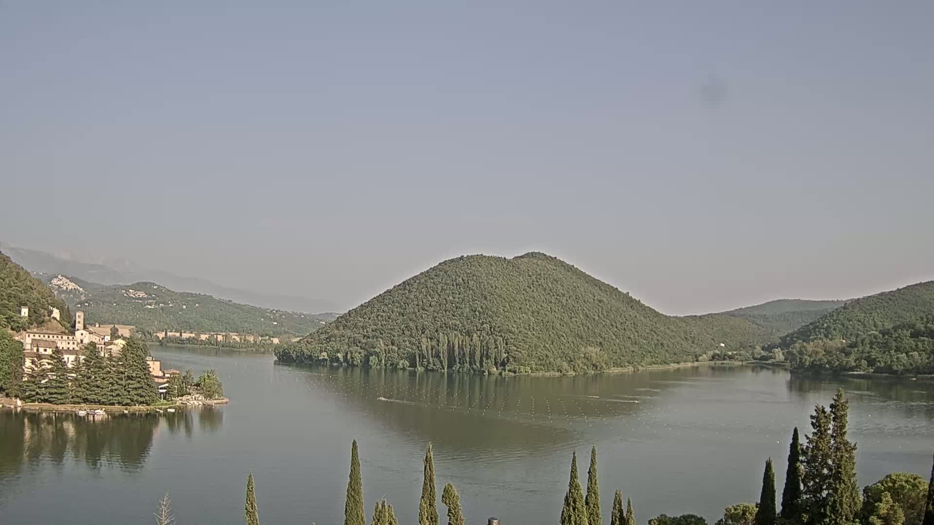 A calm lake is surrounded by green hills and a building complex nestled at the water's edge under a hazy sky.
