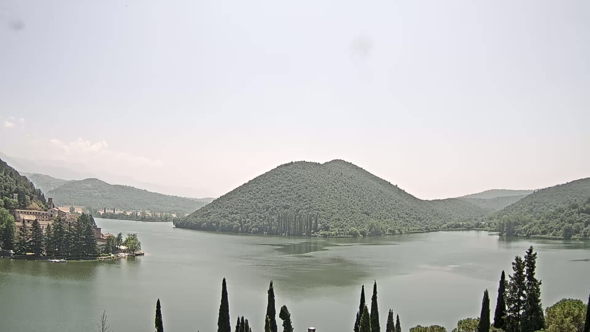 A calm lake is surrounded by lush green hills and mountains under a hazy sky.