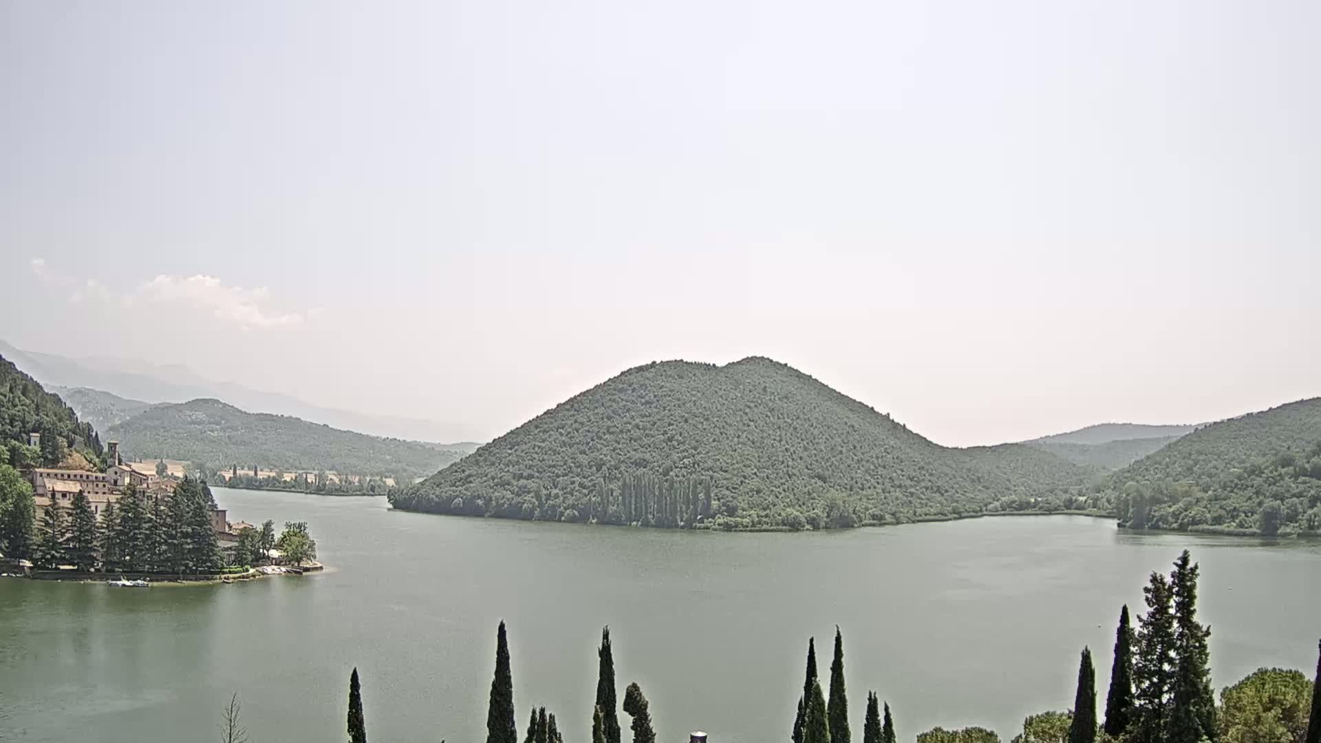 A calm lake is nestled between tree-covered hills under a hazy sky.