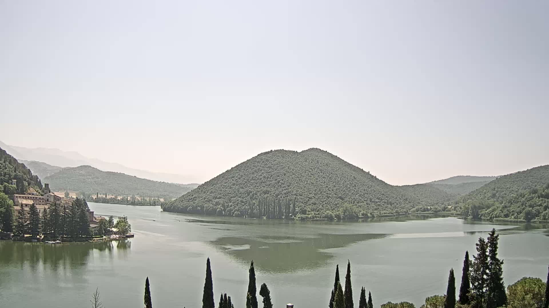 A calm lake nestled in a valley between green hills under a hazy sky.