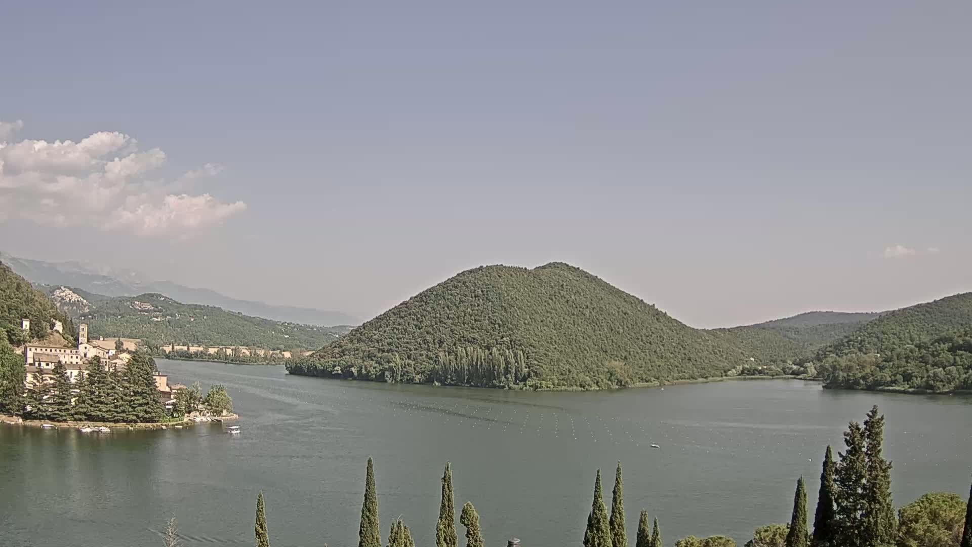 A lake nestled between tree-covered hills and mountains under a mostly clear sky with a few clouds.