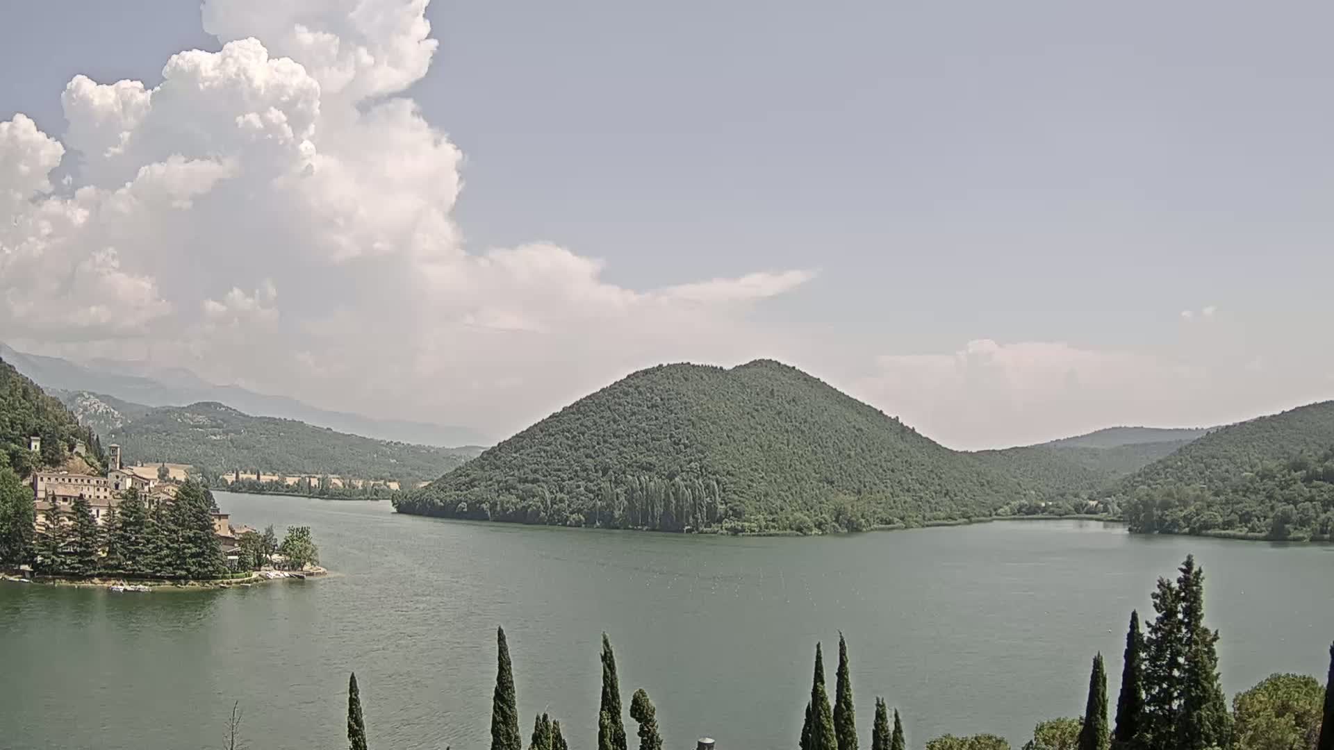 A lake is surrounded by green hills and mountains under a partly cloudy sky.