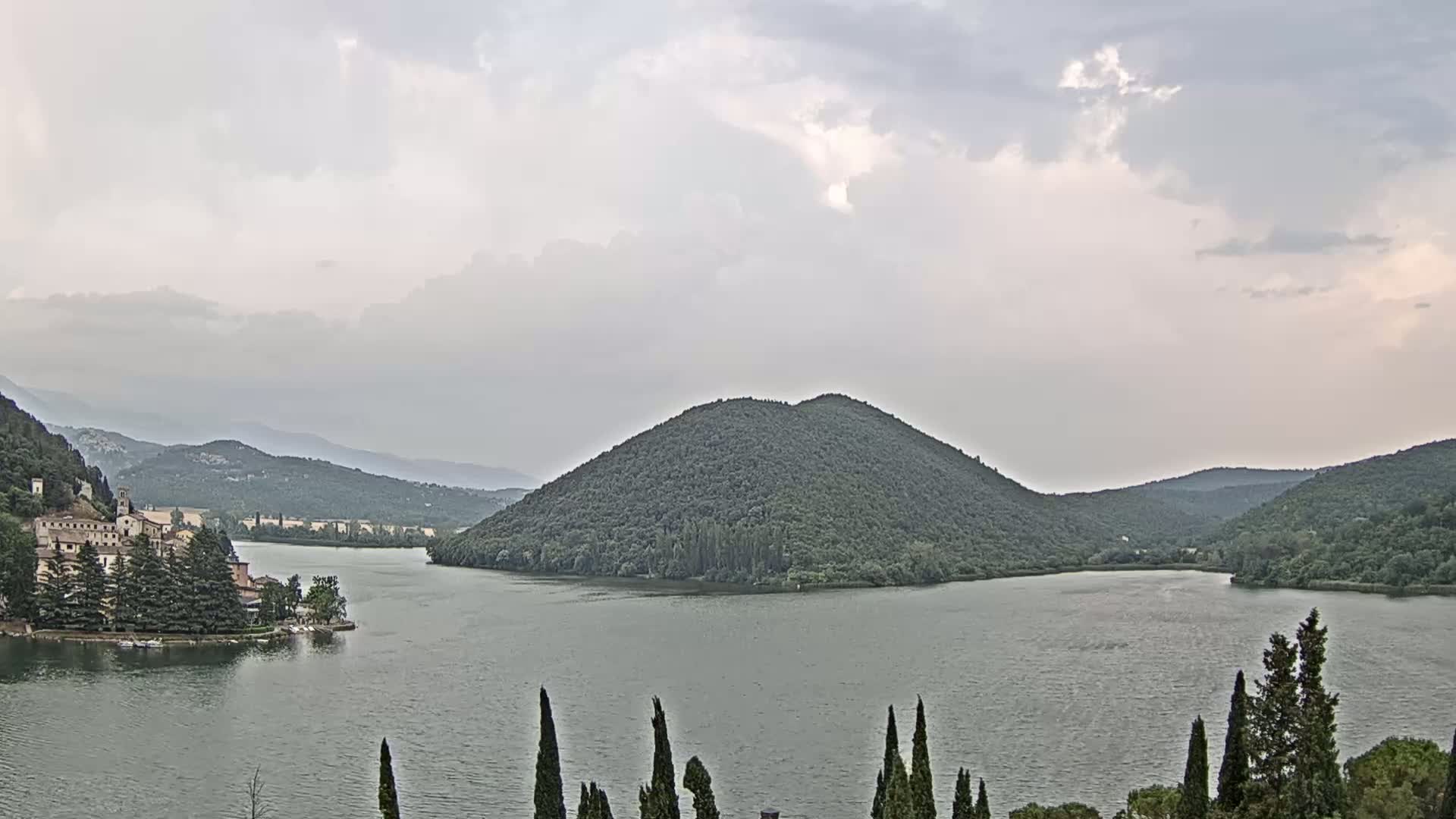 A lake is surrounded by green hills and mountains under a cloudy sky, with a village visible on the left shore.