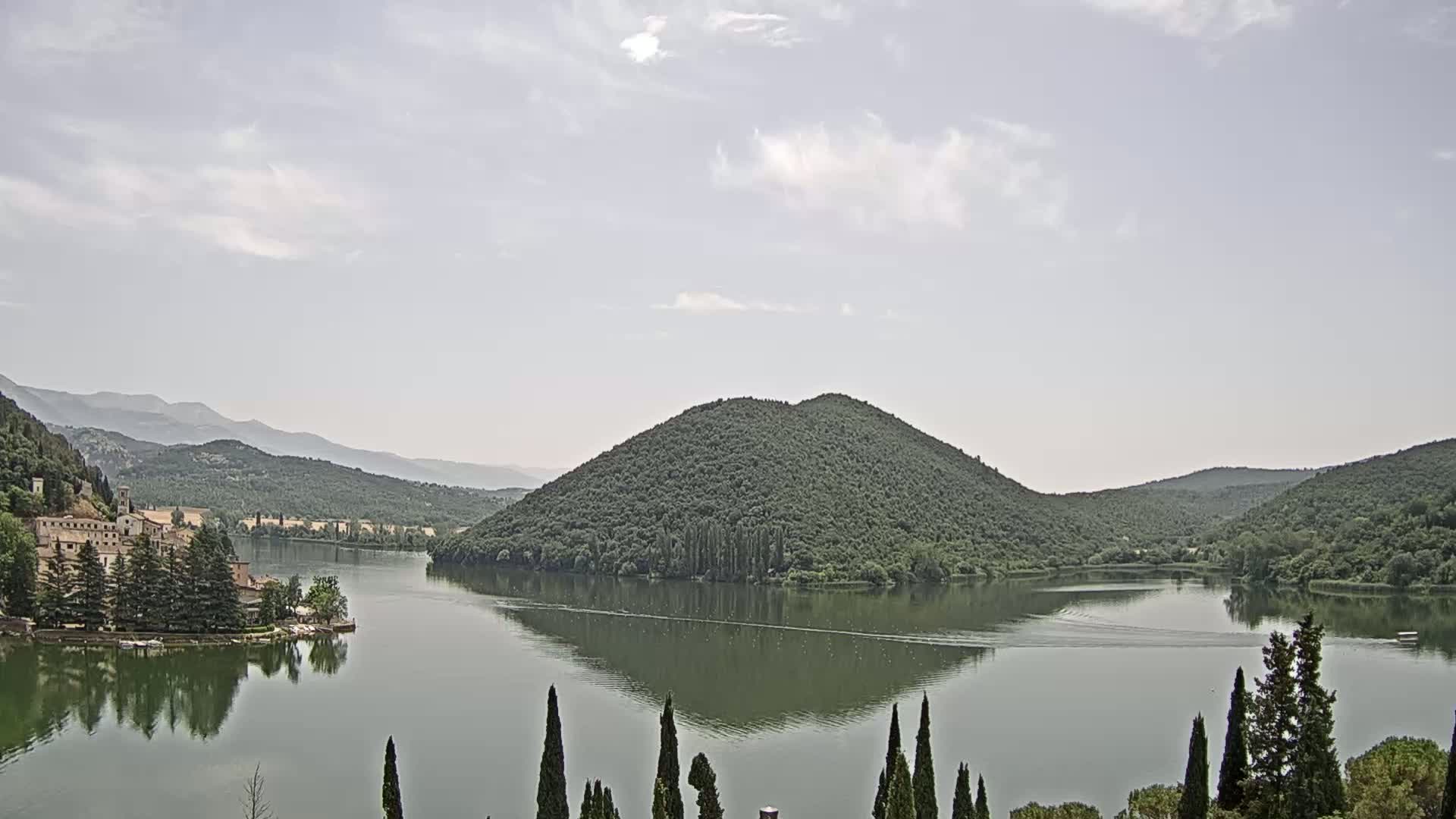 A calm lake reflects the surrounding green hills and a building complex on its shore under a partly cloudy sky.