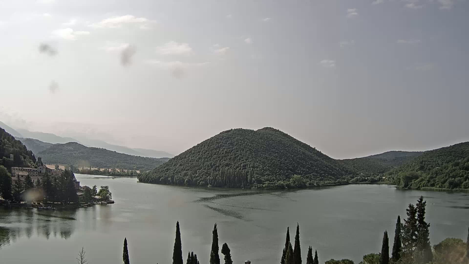 A calm lake is surrounded by green hills and mountains under a partly cloudy sky.