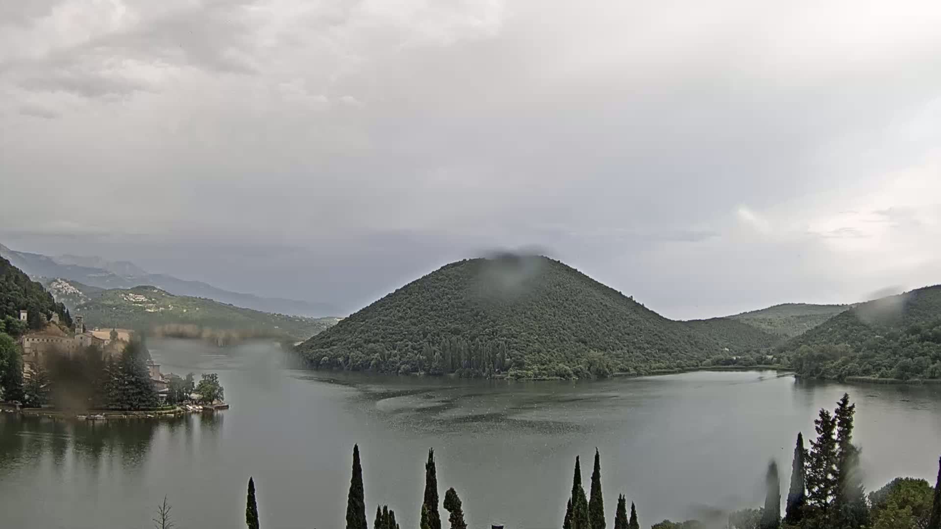 A lake surrounded by green hills and a building on the left shore is seen under a rainy, overcast sky.