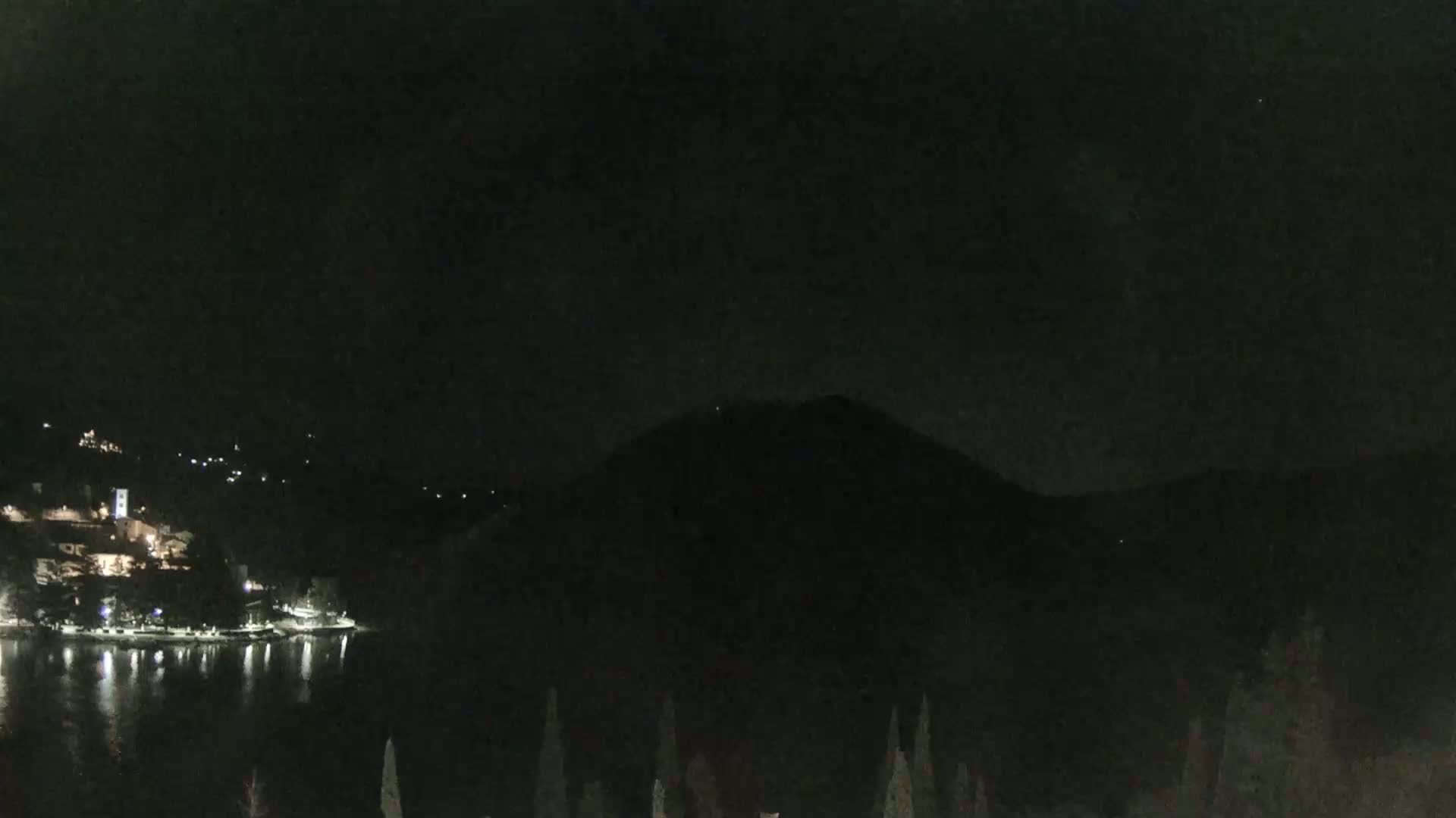 A nighttime view shows a dark mountain overlooking a calm body of water with a small, lit town on the opposite shore.