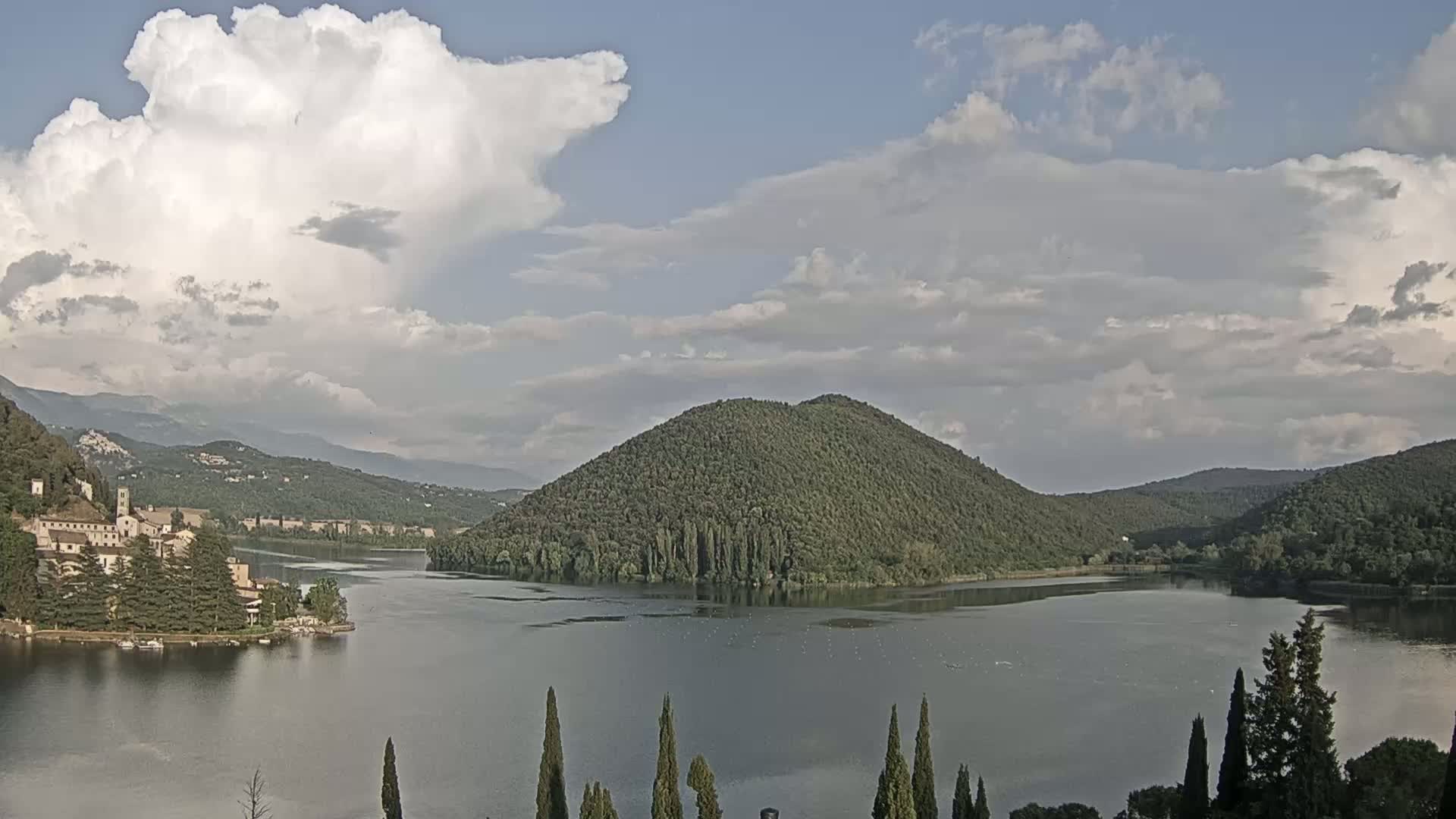 A calm lake is surrounded by green hills and a large building sits on the lakeshore under a partly cloudy sky.