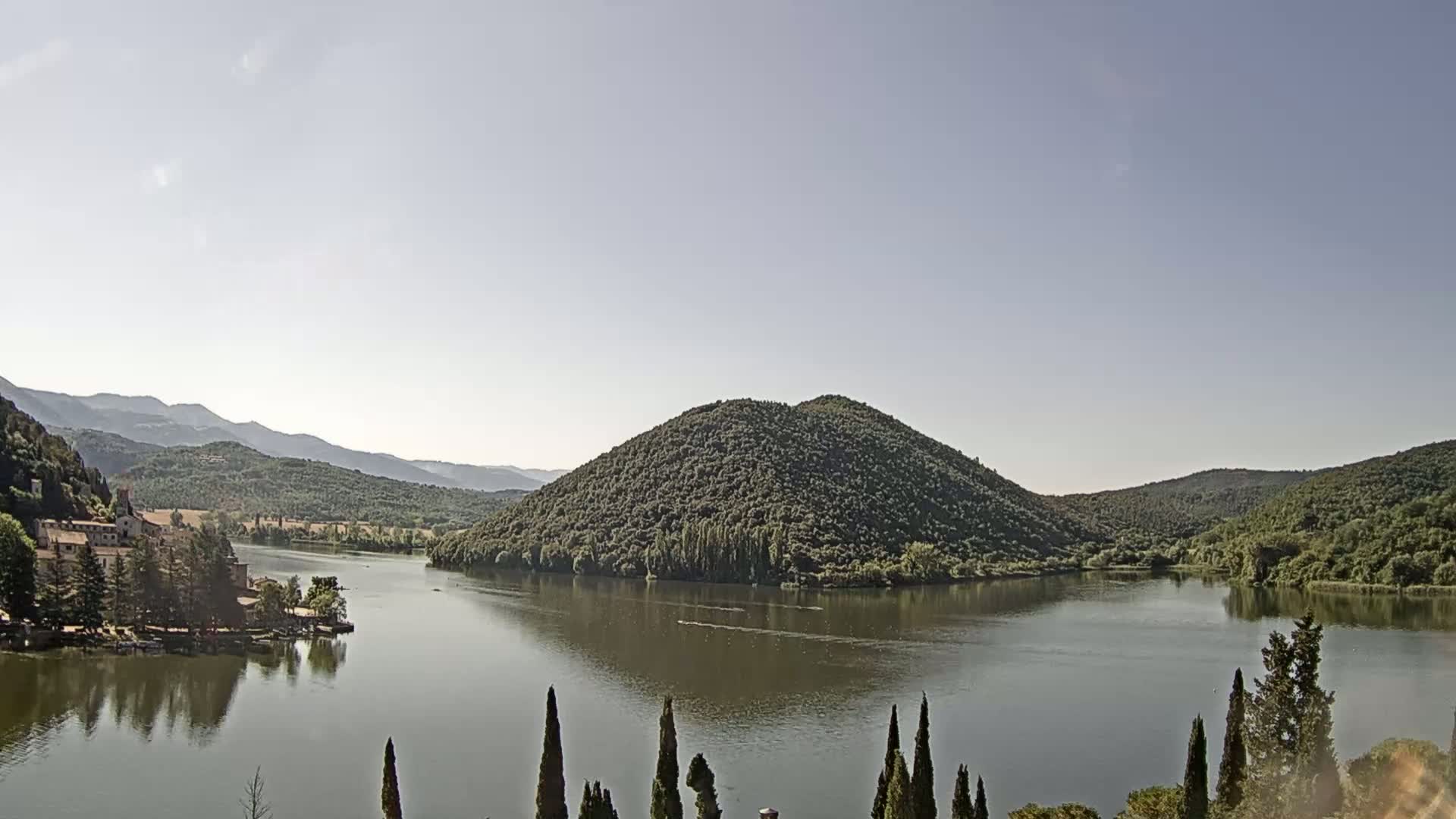 A calm lake is nestled in a valley surrounded by green hills and mountains under a clear sky.
