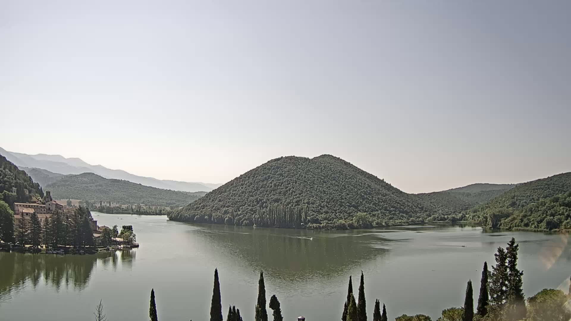 A calm lake is nestled between tree-covered hills and mountains under a hazy sky.