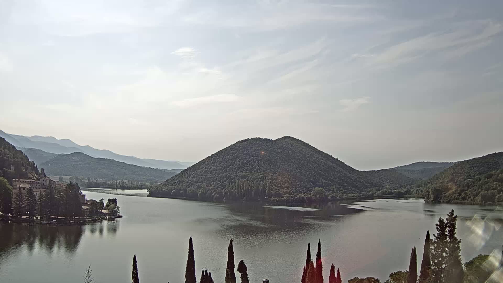 A calm lake surrounded by lush green hills and mountains under a mostly cloudy sky.