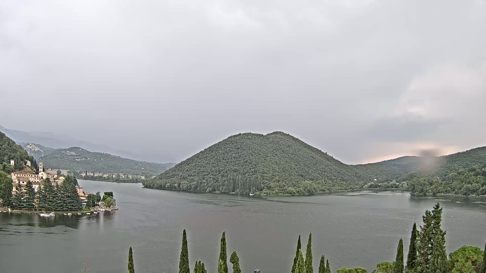 A calm lake surrounded by lush green hills and mountains under a mostly cloudy sky.