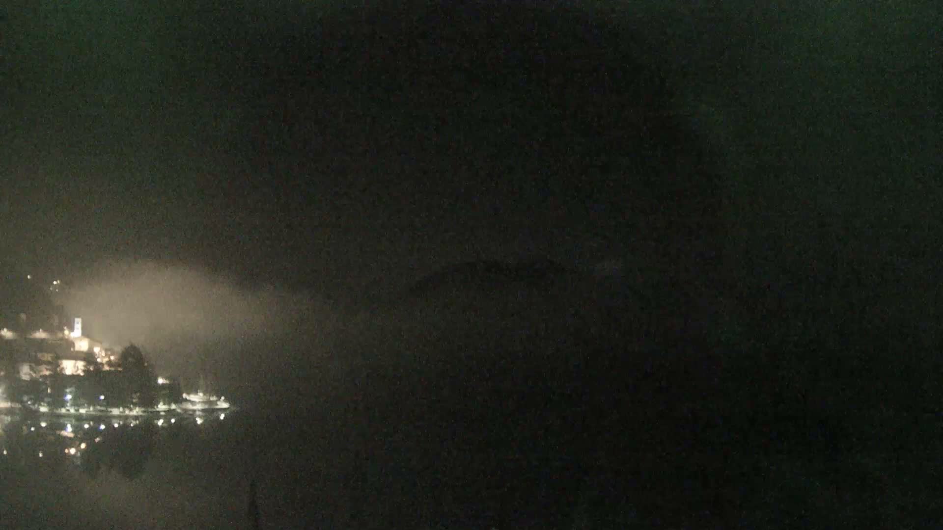 A foggy night view reveals an illuminated town nestled beside a calm body of water, with lights reflecting on the surface and faint distant mountains partially obscured by the heavy mist.