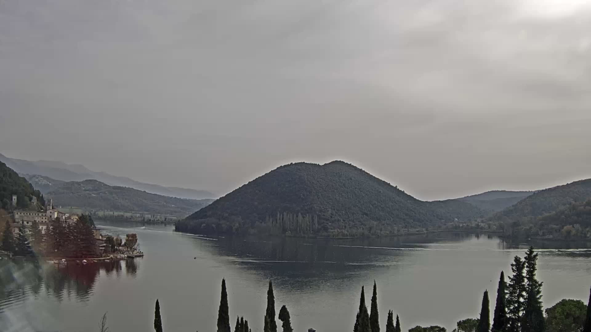Under a heavily overcast and grey sky, a tranquil lake or wide river stretches between a small waterside village backed by hills on the left and a prominent, tree-covered island in the center, with tall slender trees framing the foreground.