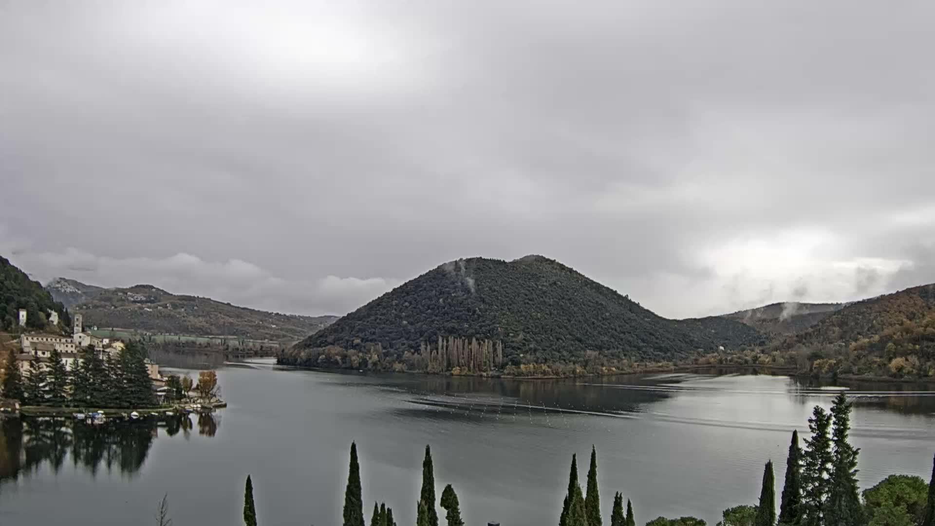Under a heavily overcast and grey sky, a tranquil lake or wide river stretches between a small waterside village backed by hills on the left and a prominent, tree-covered island in the center, with tall slender trees framing the foreground.