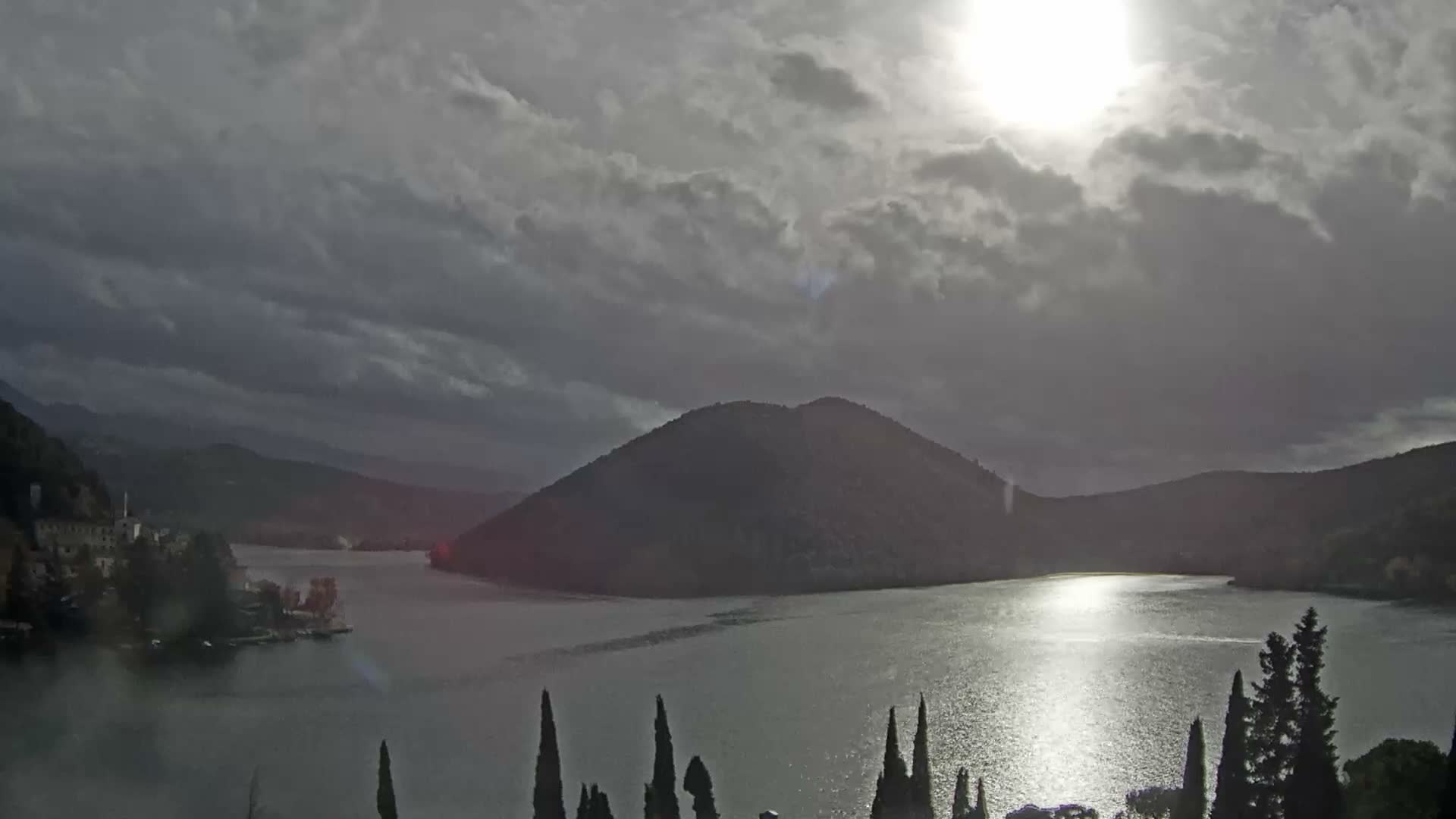 Under a heavily overcast sky with bright sun glinting off its surface, a wide lake is bordered by dark, tree-covered mountains and a small town on the left, with tall cypress trees silhouetted in the foreground.
