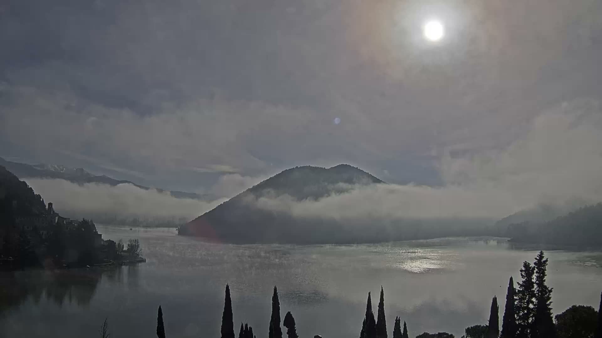 A misty lake scene features a prominent island mountain enveloped in low clouds under an overcast sky with a hazy sun, framed by dark foreground trees.