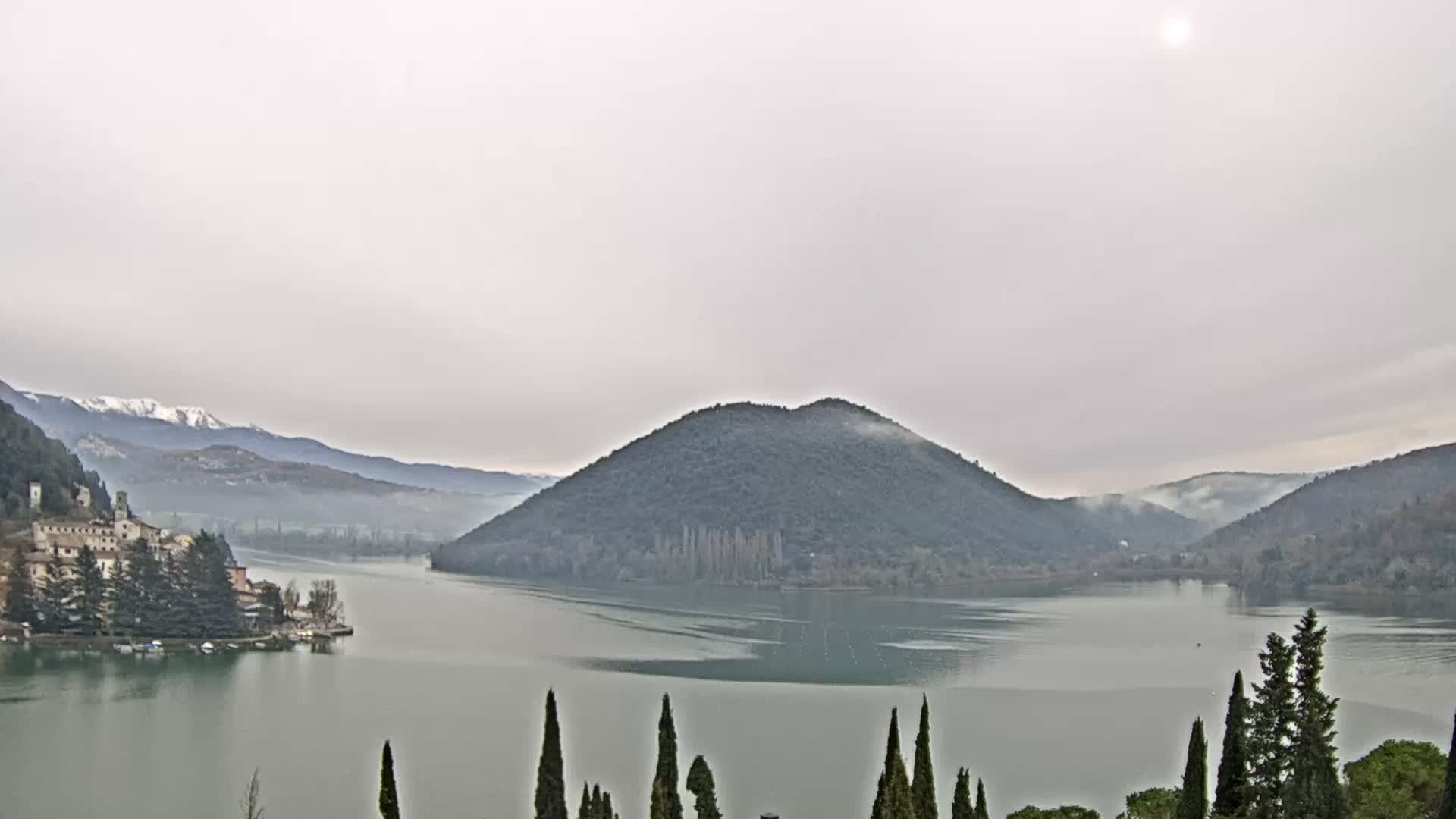 Under a uniformly overcast sky, a serene lake is framed by tree-covered hills and distant snow-capped mountains, featuring a quaint village on its left bank, a prominent wooded island in the center, and tall cypress trees in the foreground.