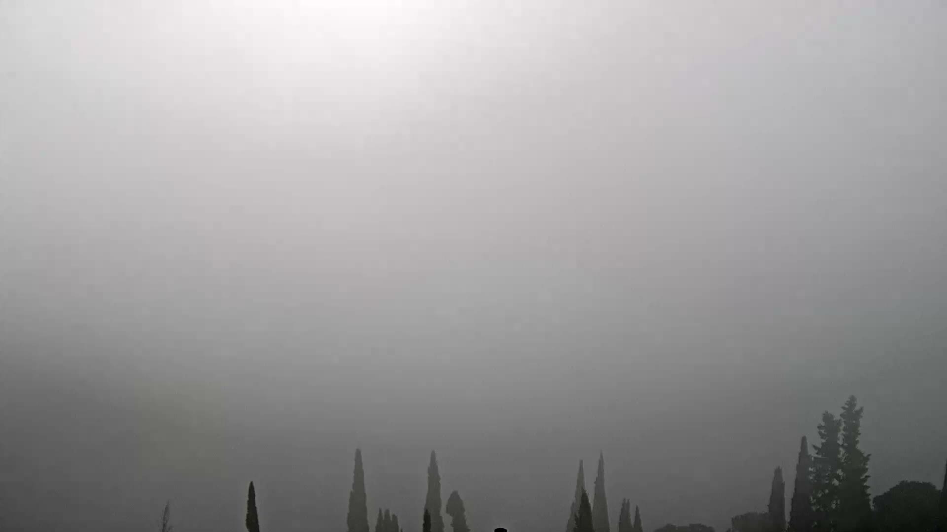 Under a uniformly overcast sky, a serene lake is framed by tree-covered hills and distant snow-capped mountains, featuring a quaint village on its left bank, a prominent wooded island in the center, and tall cypress trees in the foreground.