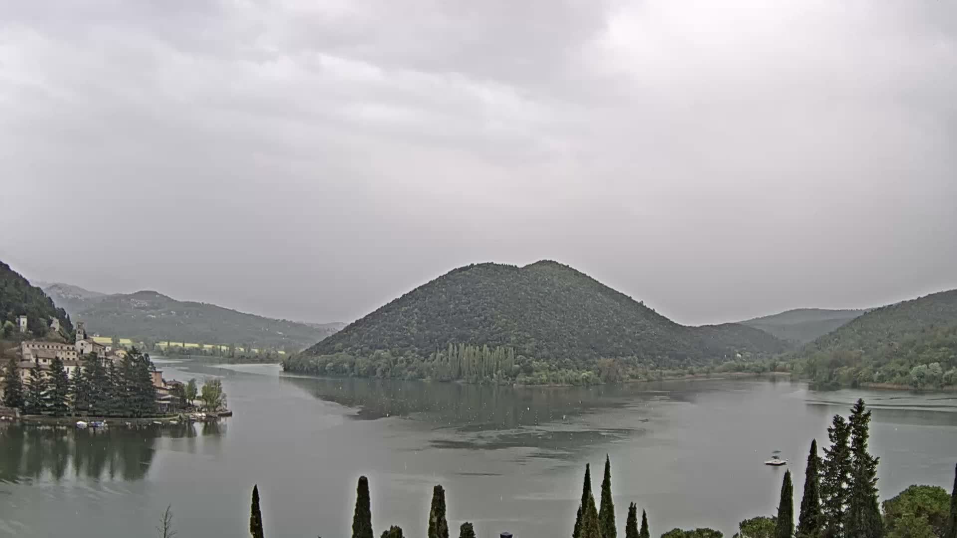 Under a uniformly overcast sky, a serene lake is framed by tree-covered hills and distant snow-capped mountains, featuring a quaint village on its left bank, a prominent wooded island in the center, and tall cypress trees in the foreground.