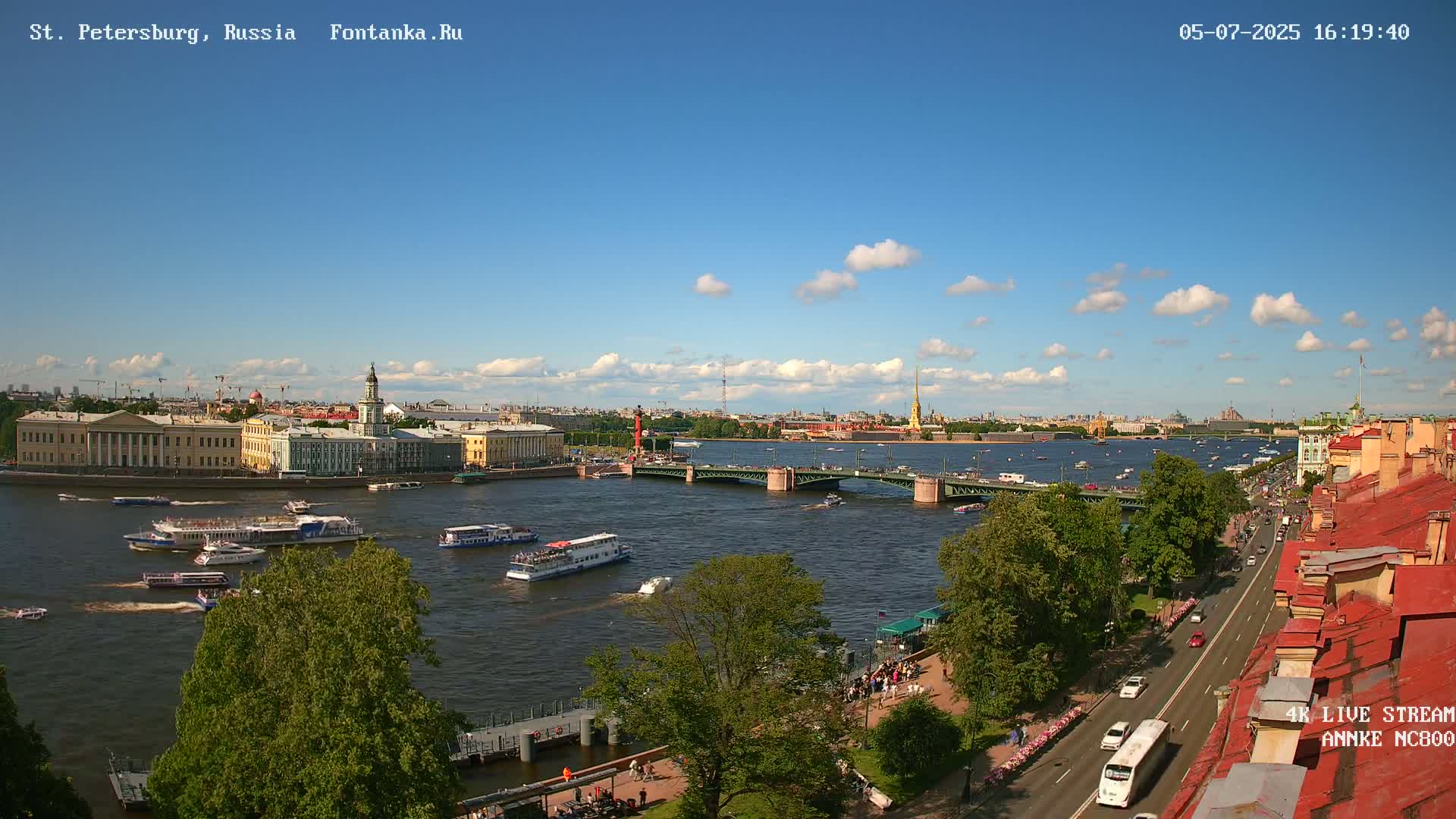 A sunny day shows a river with several tour boats, a bridge, and city buildings along the water's edge, all under a partly cloudy sky.