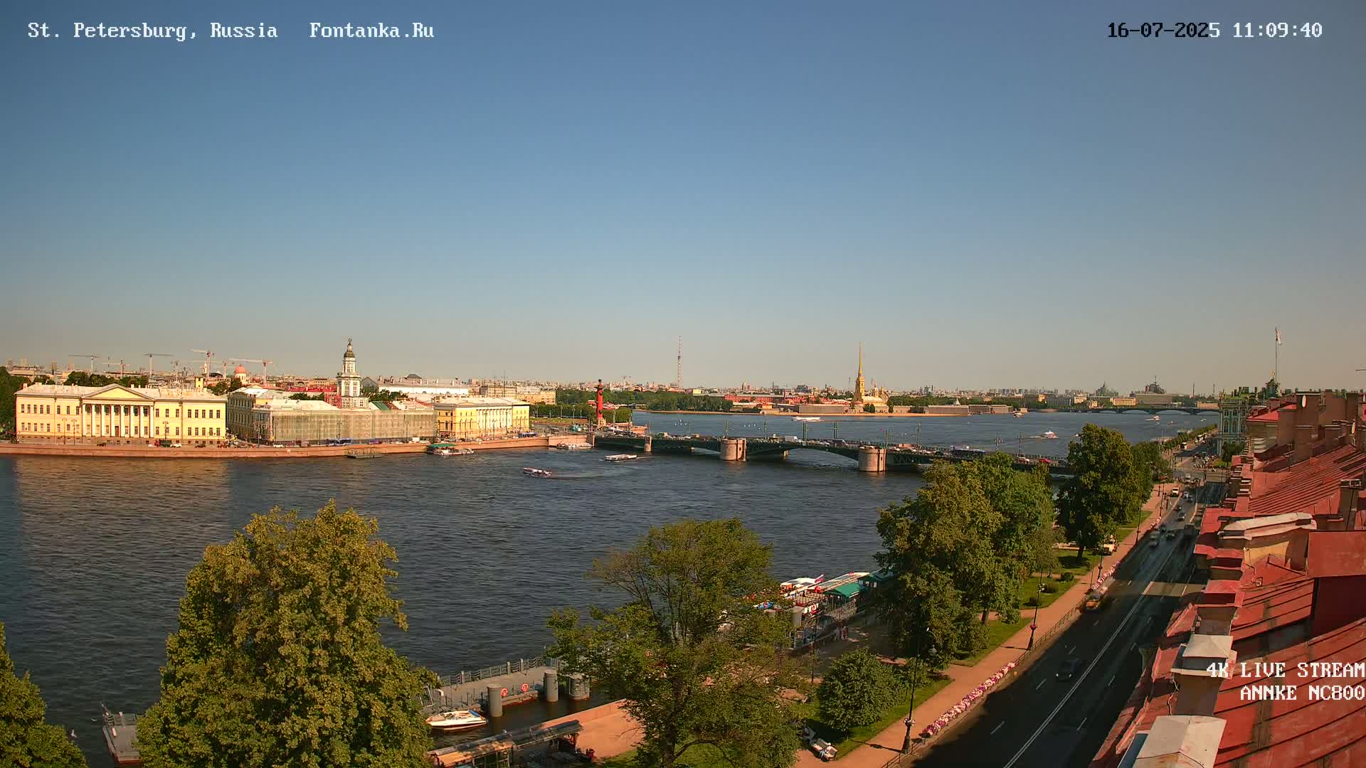 A clear, sunny day shows a river with a bridge, buildings, and green trees lining the riverbank.