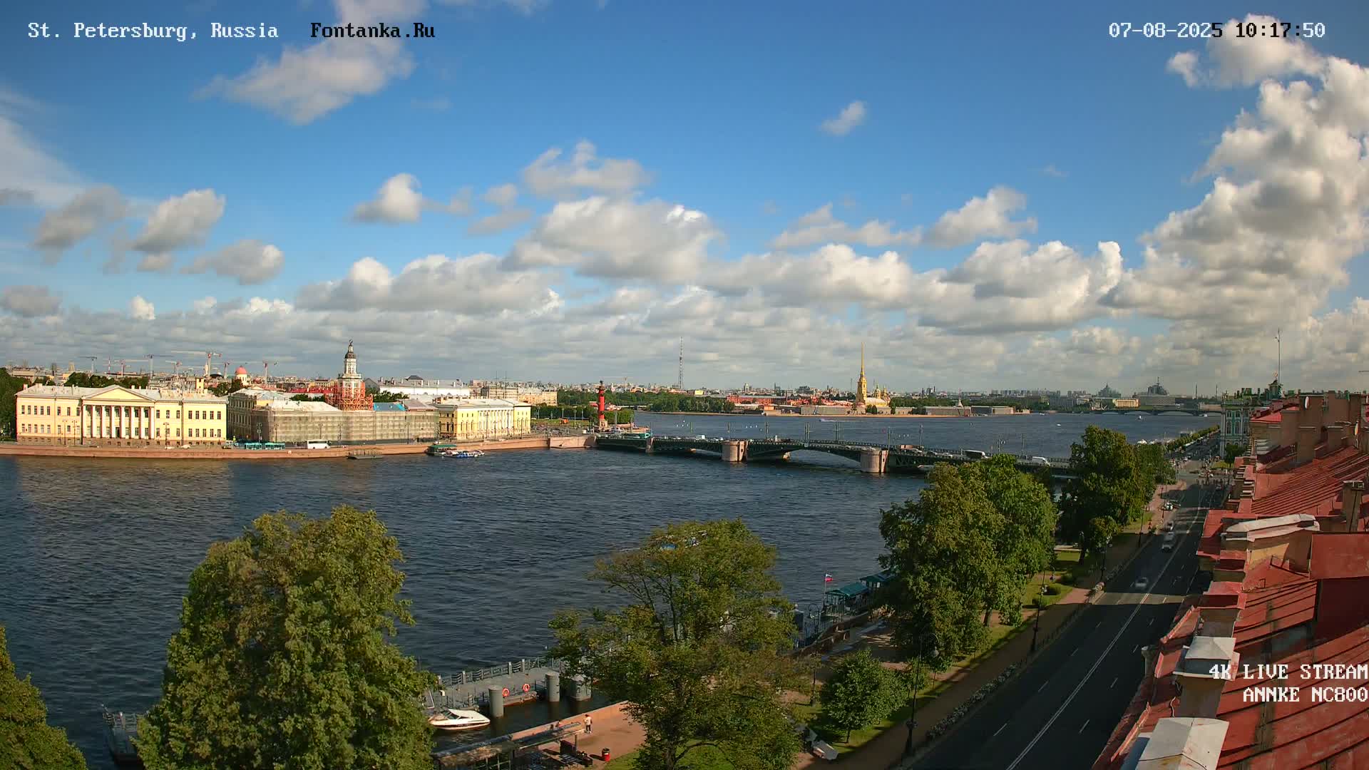 A partly cloudy sky overlooks a river scene with a bridge, buildings, and lush green trees.