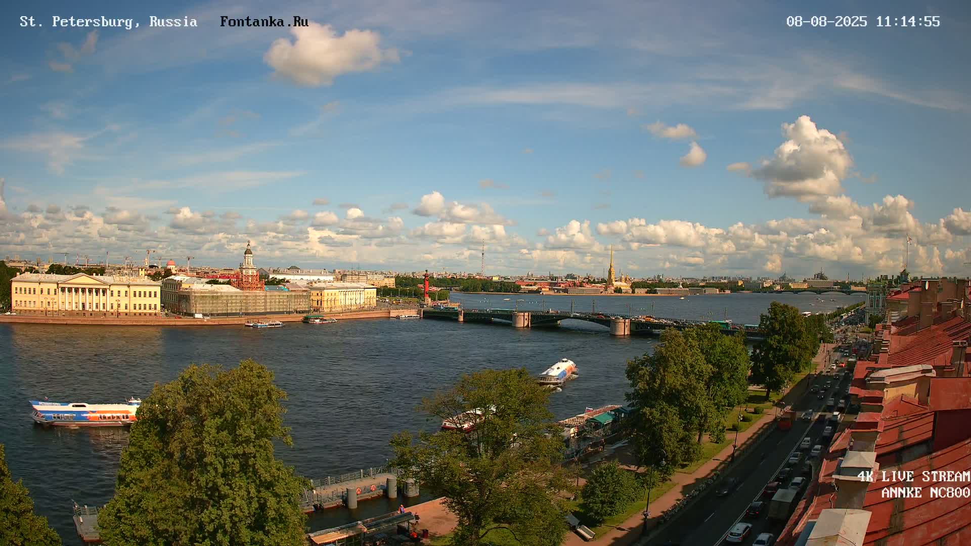 A sunny day with partly cloudy skies shows a city river scene with buildings, bridges, boats, and traffic along a waterfront road.