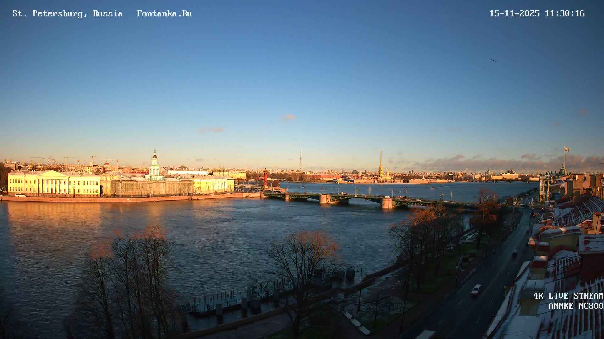 An expansive outdoor view captures a city's waterfront with a wide river, multiple bridges, and historical buildings glowing under low golden sunlight beneath a clear blue sky.