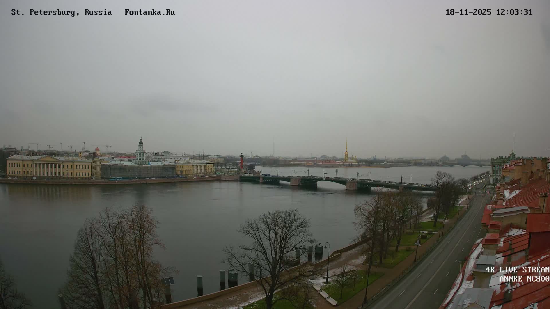 An expansive outdoor view captures a city's waterfront with a wide river, multiple bridges, and historical buildings glowing under low golden sunlight beneath a clear blue sky.