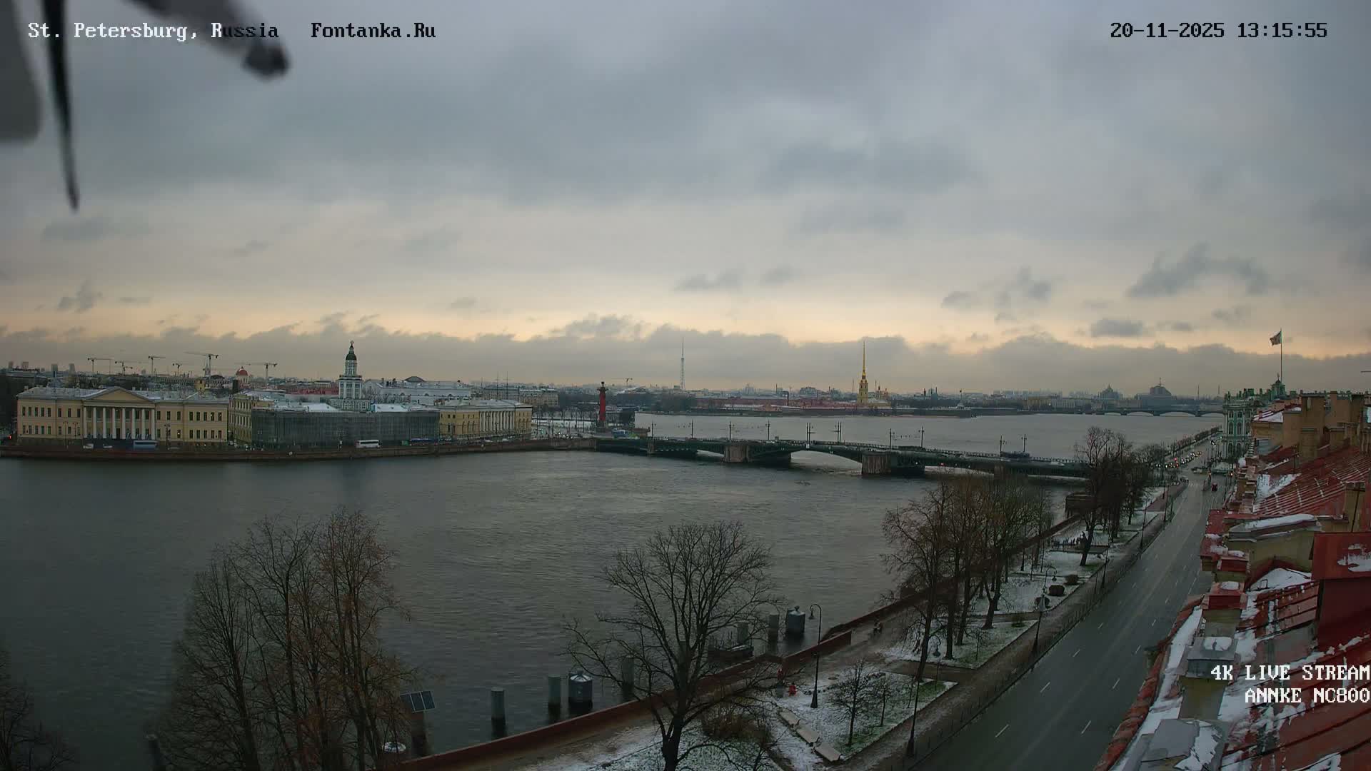 A wide river flows through a city on an overcast winter day, with bridges connecting banks lined by historic buildings and a snow-dusted promenade, under a sky with hints of twilight colors.