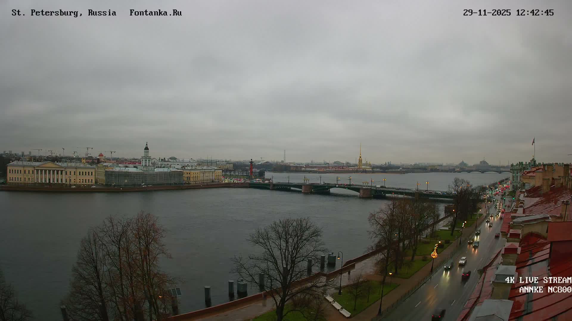 A wide river flows through a city under a gloomy, overcast sky, flanked by historic buildings and a busy wet road with vehicle traffic.