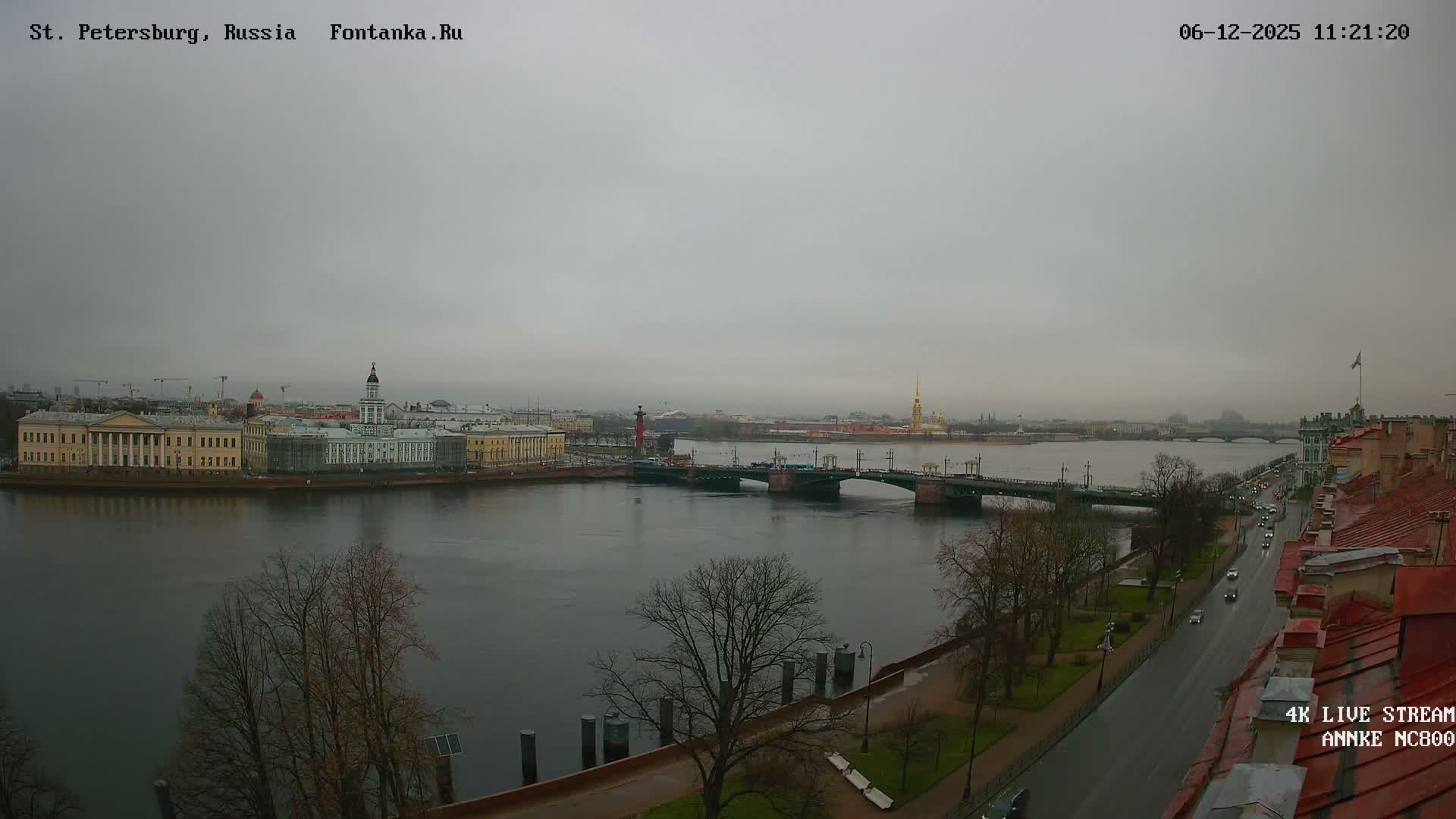 A wide, gray river flows through a historic European city under a heavy overcast sky, flanked by grand buildings, a distant golden spire, a bridge, and a busy riverside road with sparse, bare trees, indicative of cool, damp weather.