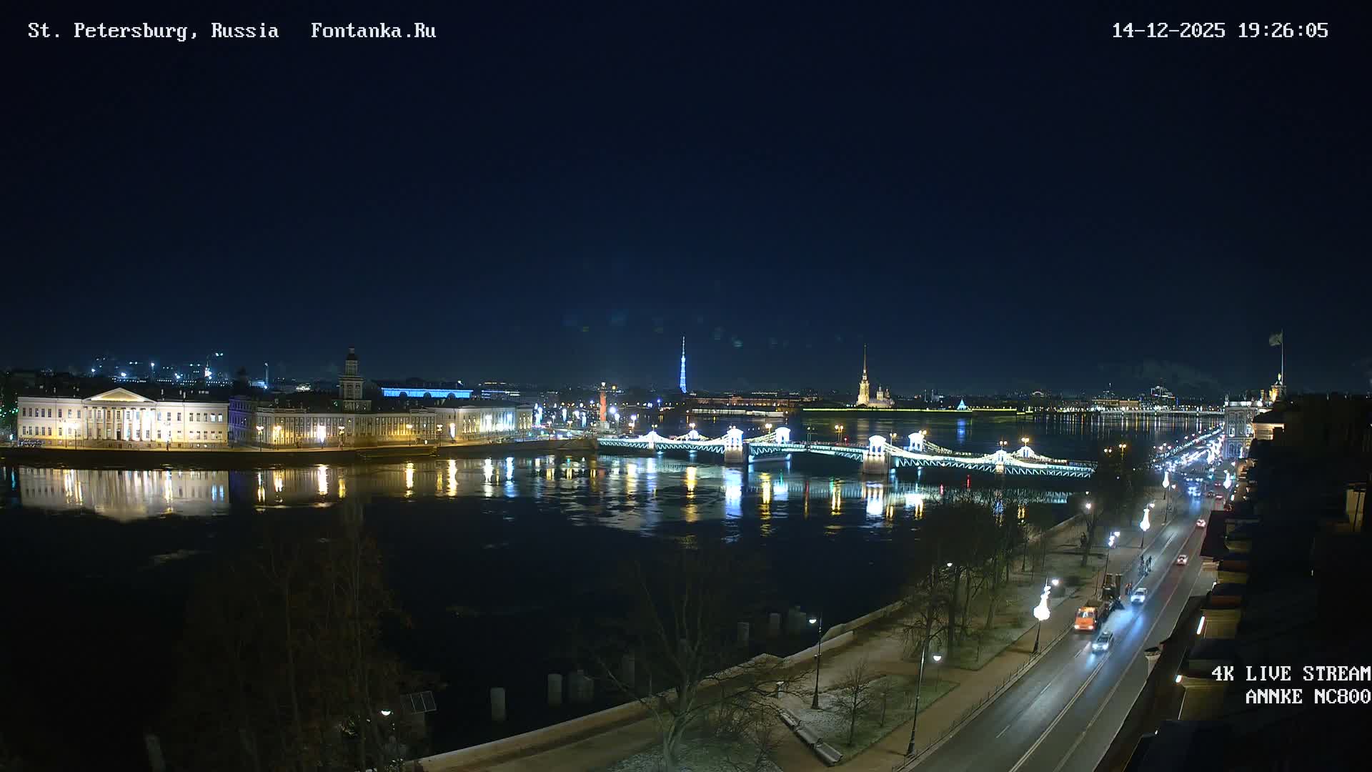 A wide, gray river flows through a historic European city under a heavy overcast sky, flanked by grand buildings, a distant golden spire, a bridge, and a busy riverside road with sparse, bare trees, indicative of cool, damp weather.