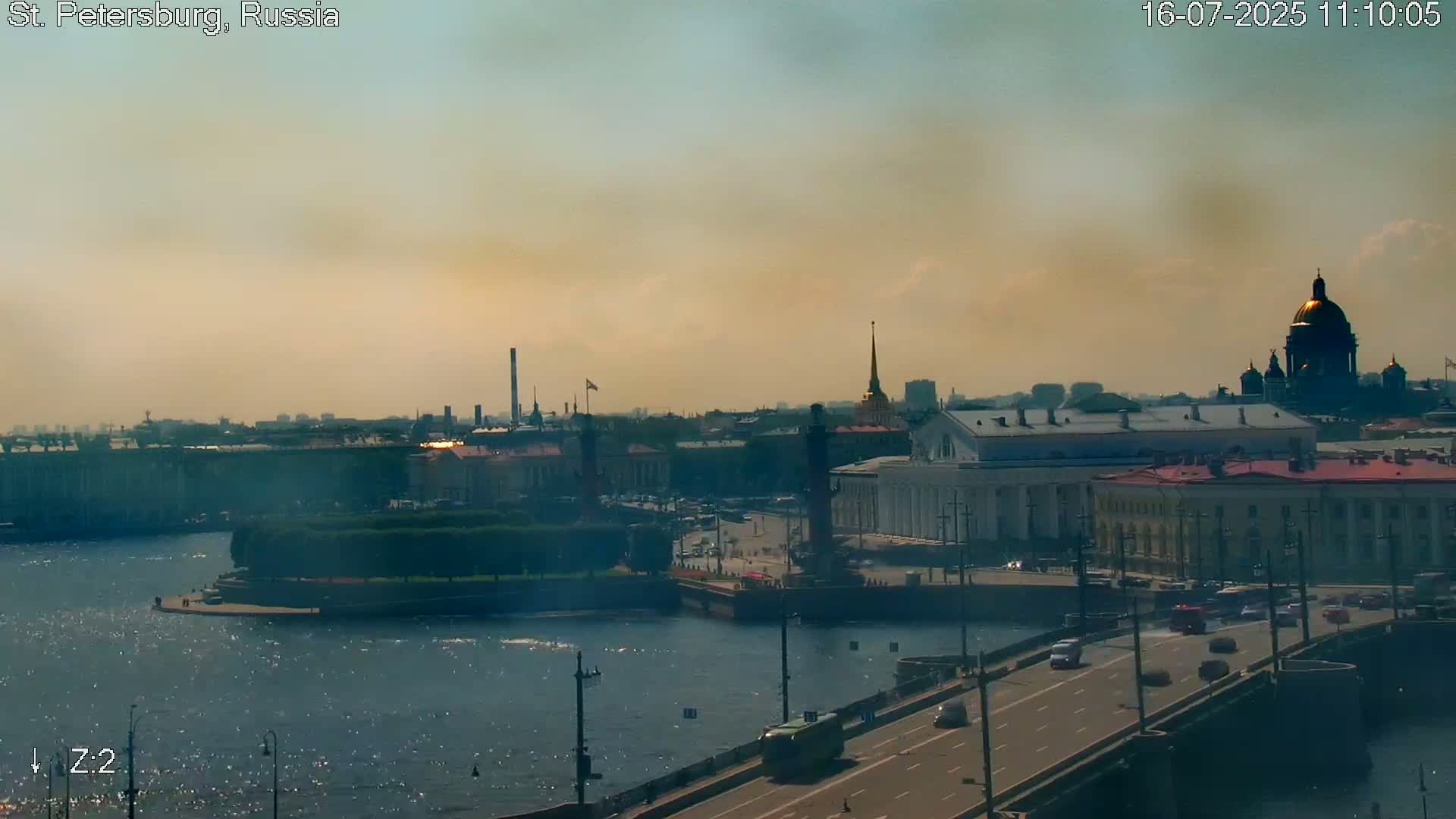 A hazy, sunny cityscape overlooks a river with a bridge carrying cars, featuring prominent buildings and a golden-domed cathedral.
